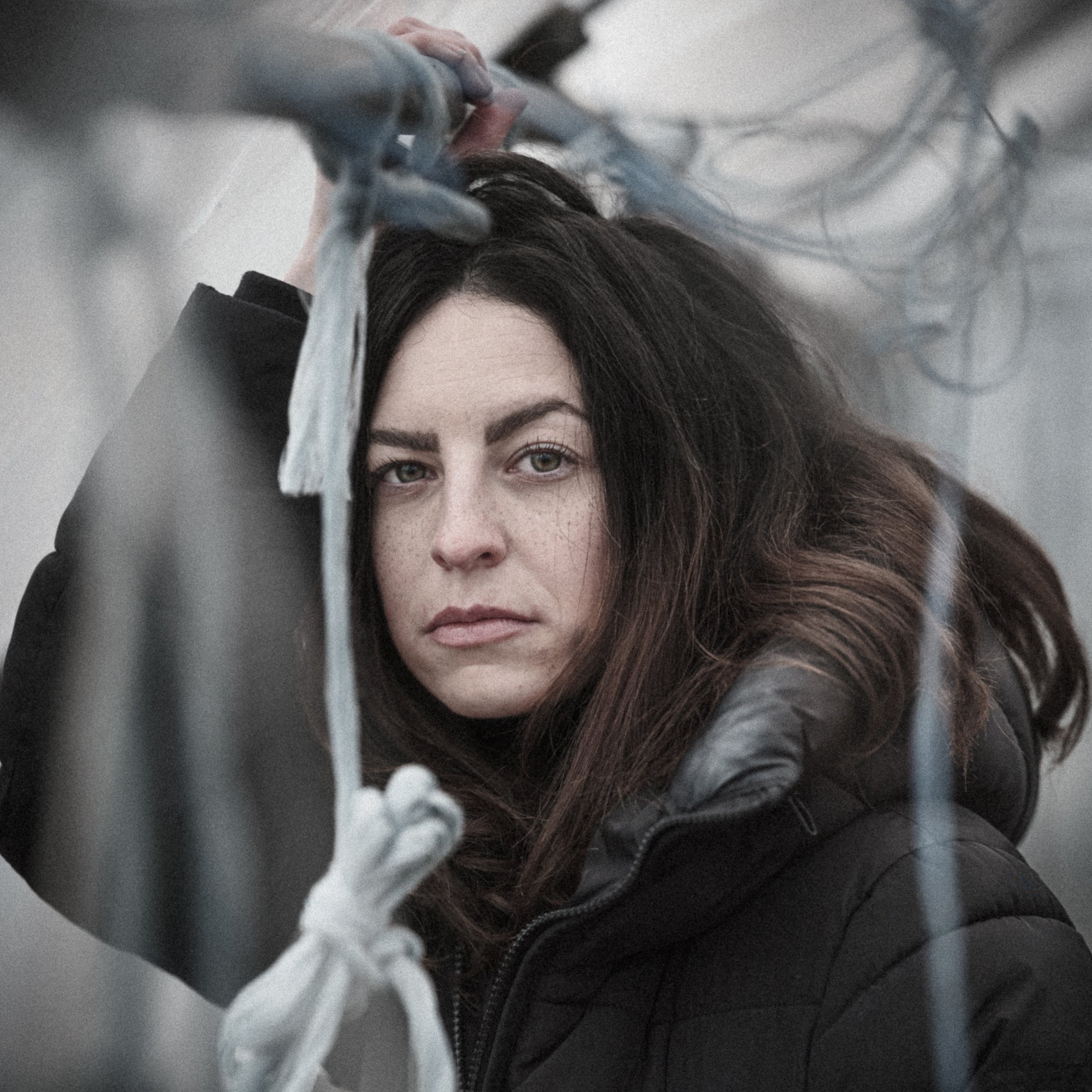 Close-up of a woman with dark brown hair and brown eyes, holding onto a metallic railing with her hand, with a blurred background.