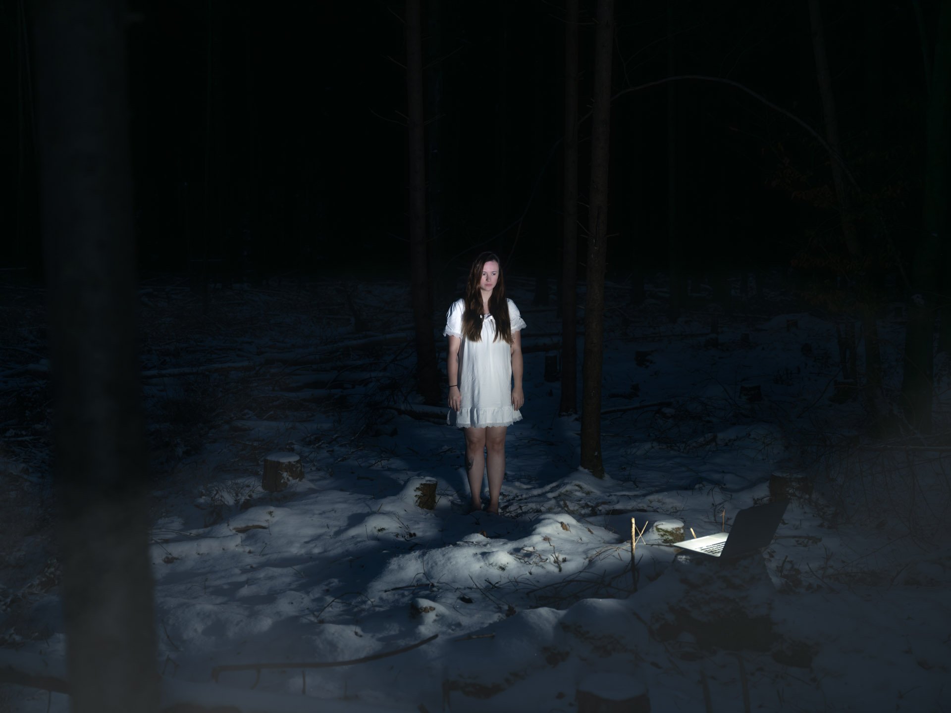 A young woman with long brown hair wearing a white dress standing in a dark, snow-covered forest at night, illuminated by a strong light source.