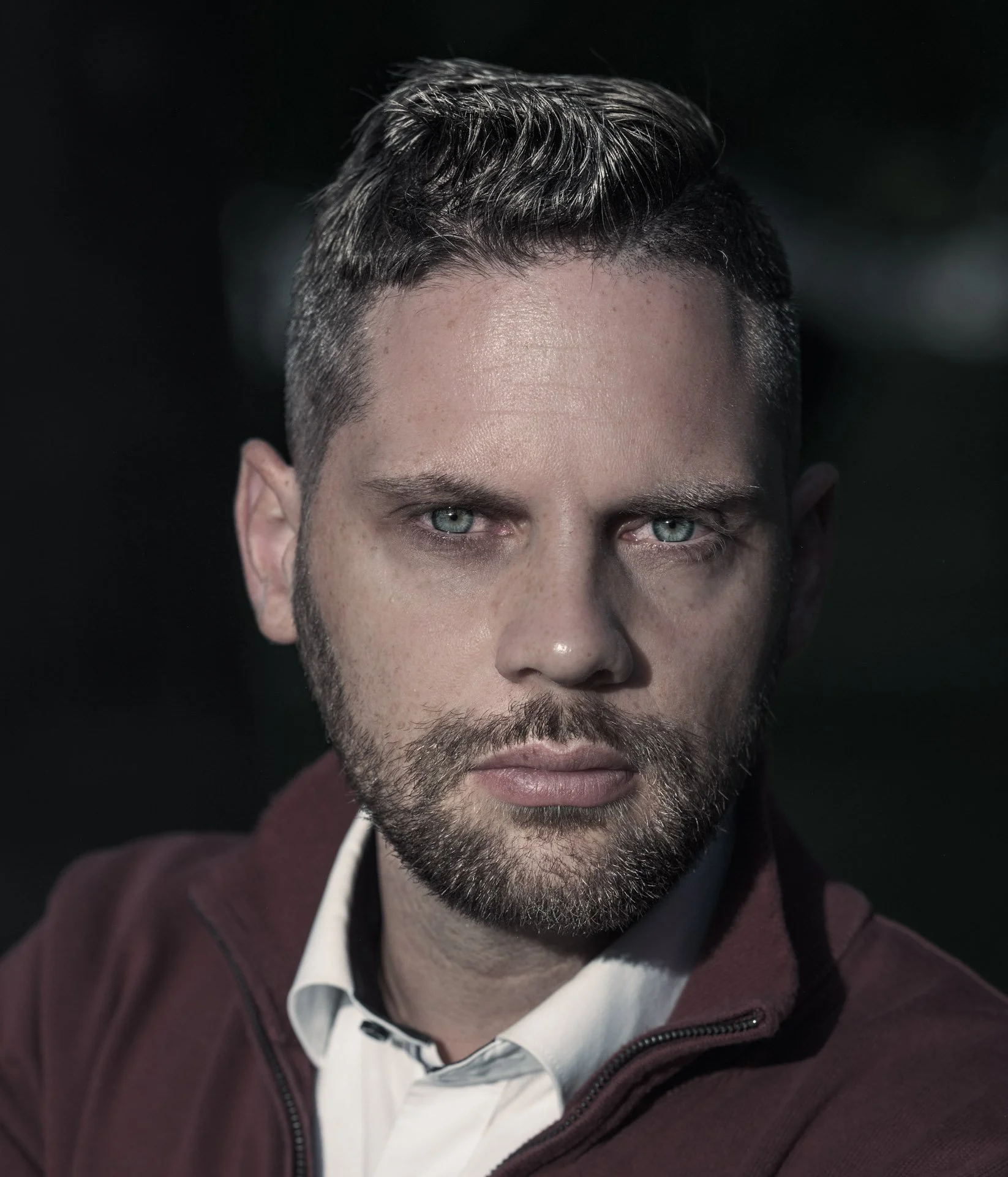 A close-up portrait of a man with green eyes, dark hair, and a beard, wearing a white shirt and maroon jacket, against a dark background.