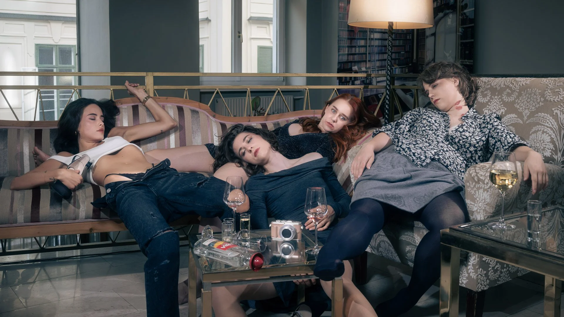 Four busted women sitting and lying on couches with empty wine glasses and bottles, in a living room setting.