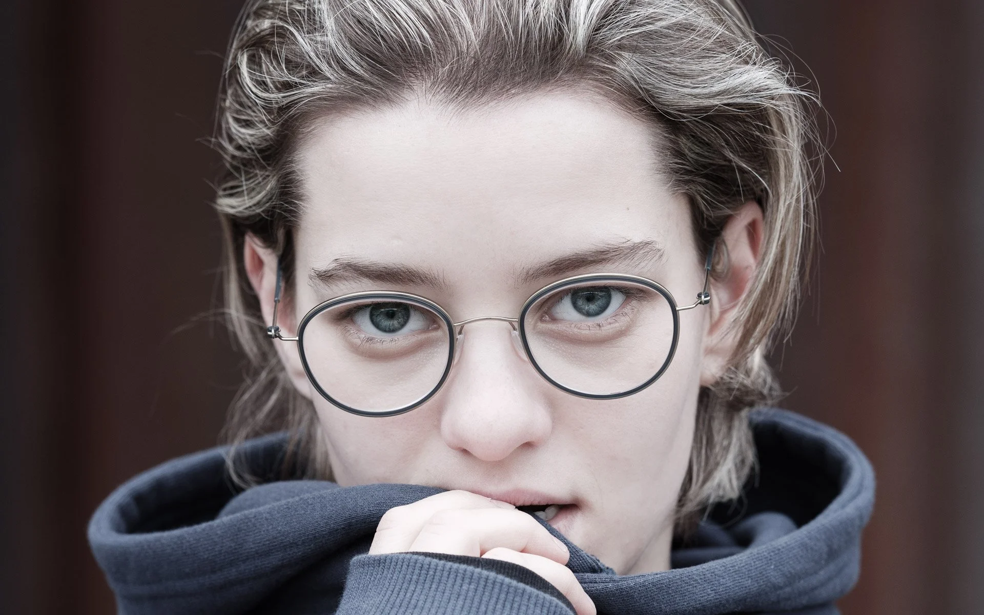A young person with light skin, light-colored hair, and glasses looking directly at the camera with one hand near their mouth, biting in a dark hoodie.