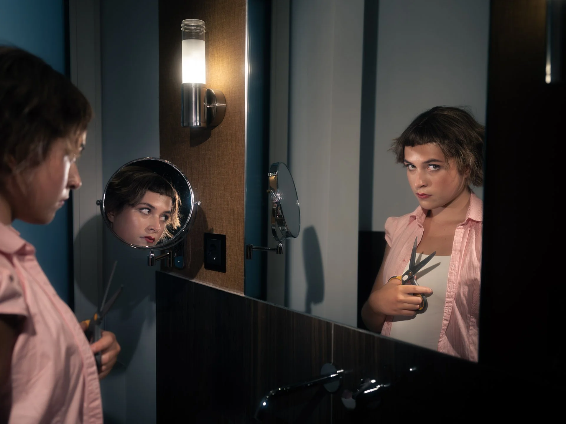 A young woman with short brown hair and wearing a pink shirt holds a pair of scissors in front of a mirror in a bathroom, looking directly at her reflection with a serious expression.