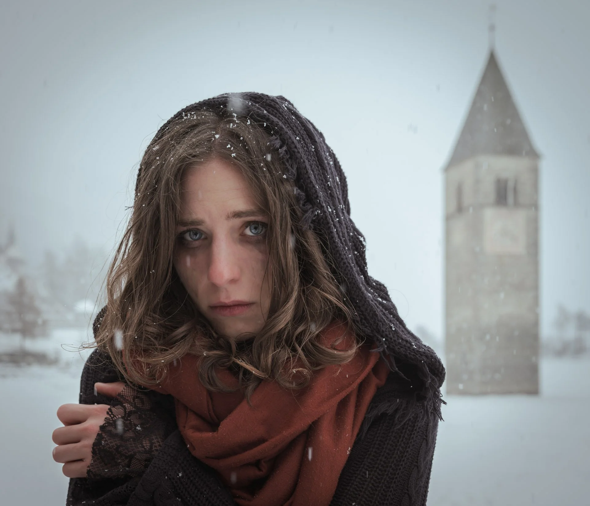 A woman with brown hair and blue eyes, wearing a black knit hat and a rust-colored scarf, standing outdoors in a snowy setting with a church steeple in the background.