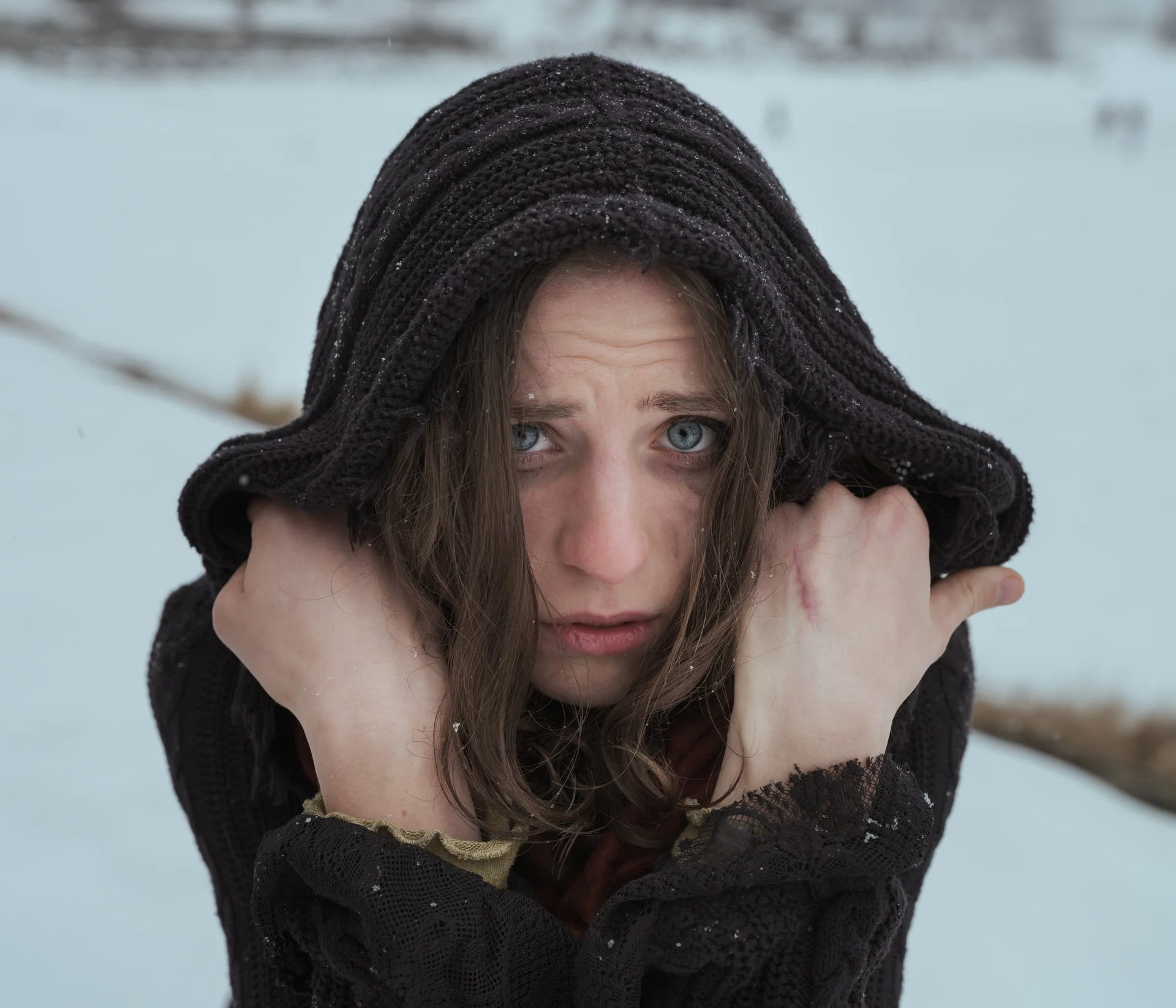 Close-up of a woman with blue eyes and brown hair, wearing a black knit hooded sweater in a snowy outdoor setting.