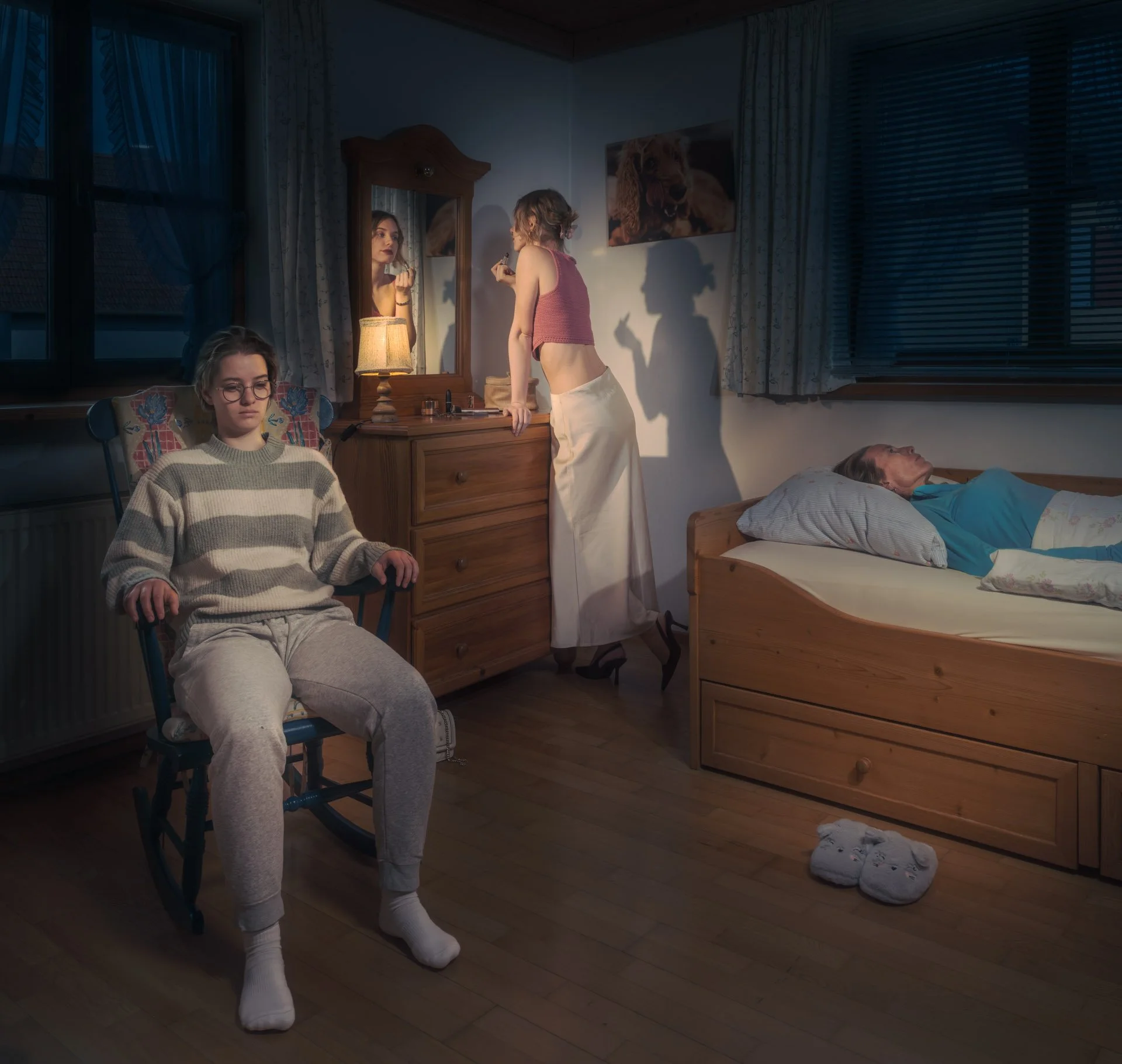 A young woman applying makeup at a mirror in a dimly lit bedroom. An older woman is lying in bed, and a young woman with glasses sits in a chair, wearing cozy loungewear. There is a dog painting on the wall, a bedside lamp, and slippers on the wooden floor.
