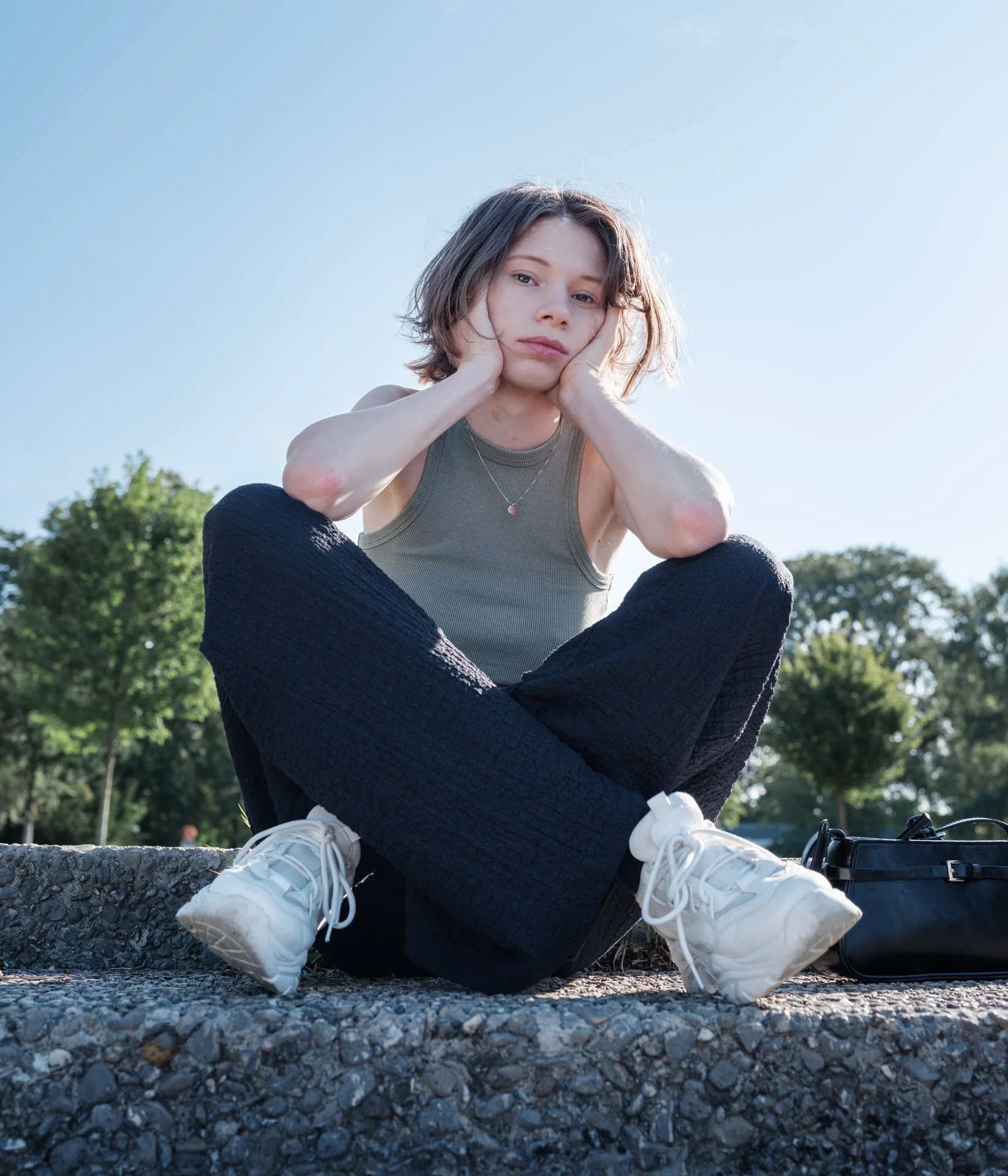 Young woman sitting outdoors on a stone surface, resting her head in her hands, wearing a gray tank top and black pants, with white sneakers. She has short, wavy brown hair and a necklace. The background features trees and a clear sky.