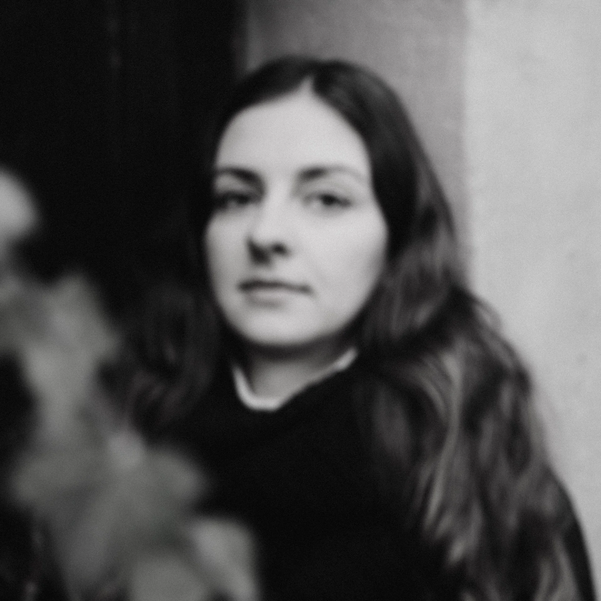 Black and white, blurred, portrait of a woman with long wavy hair, looking at the camera with a neutral expression.