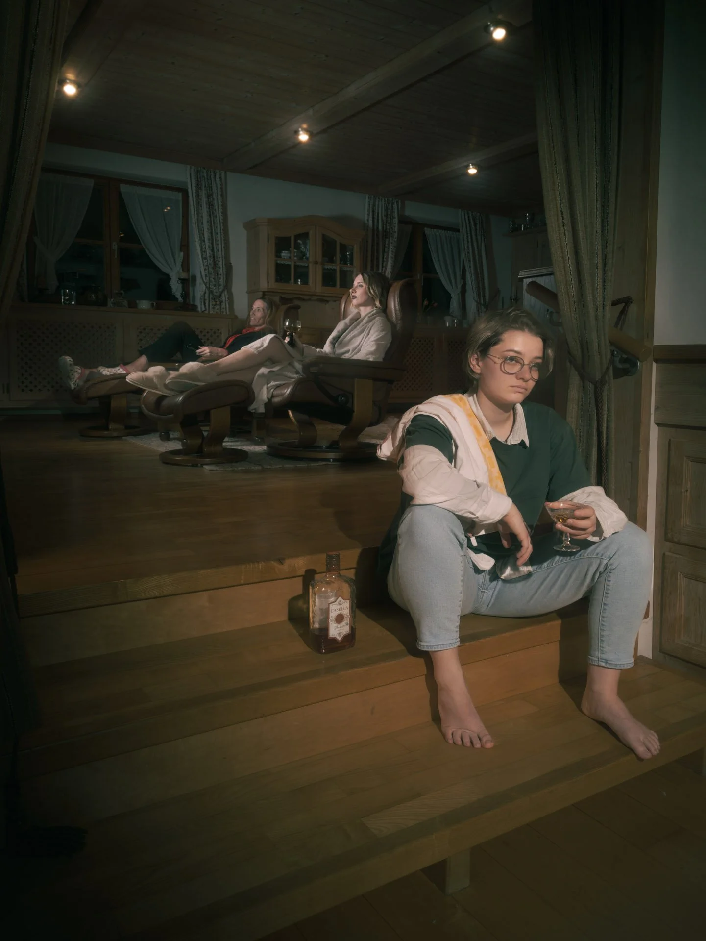A woman sitting barefoot on wooden steps holding a glass of wine in a cozy, dimly lit room, with two women lounging on recliners in the background, one holding a glass of wine, and a bottle of wine on the floor in the foreground.