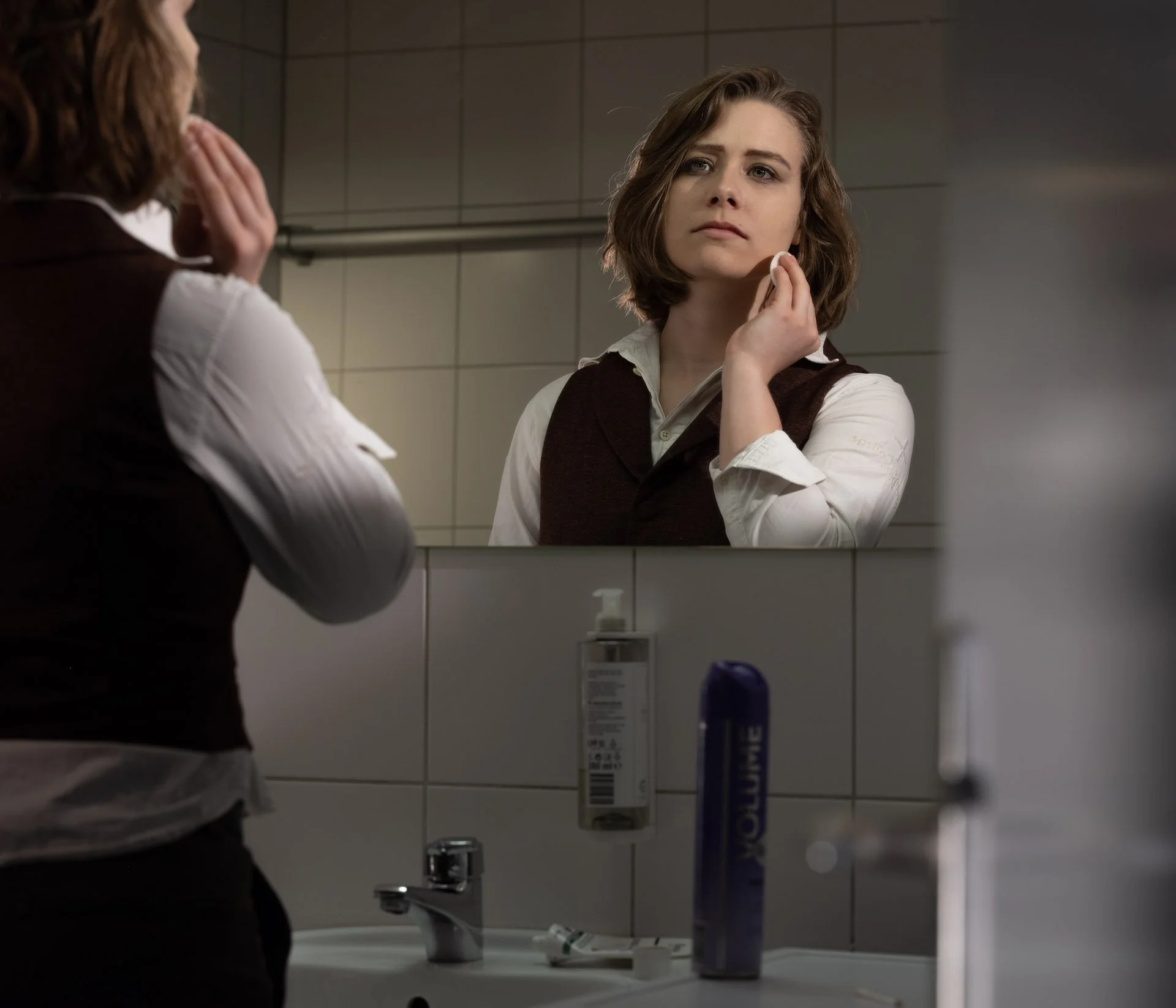 A woman with short brown hair looking into a bathroom mirror and touching her face.