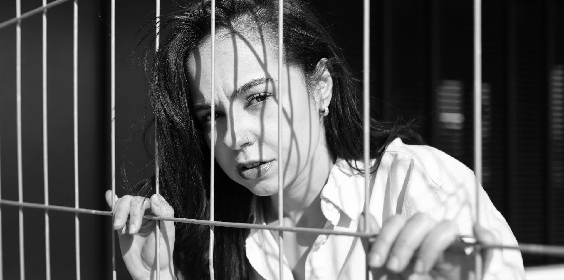 Black and white photo of a woman with dark hair touching a metal fence, looking through it with a serious expression.