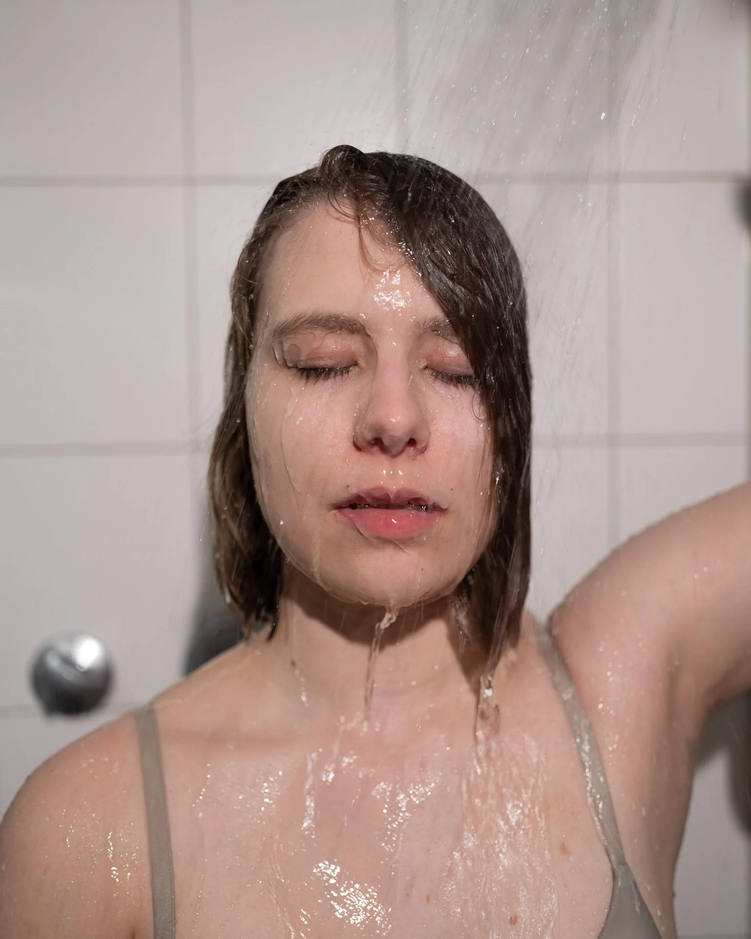 A woman with wet hair and closed eyes, standing under a shower with water pouring over her face and body in a tiled bathroom
