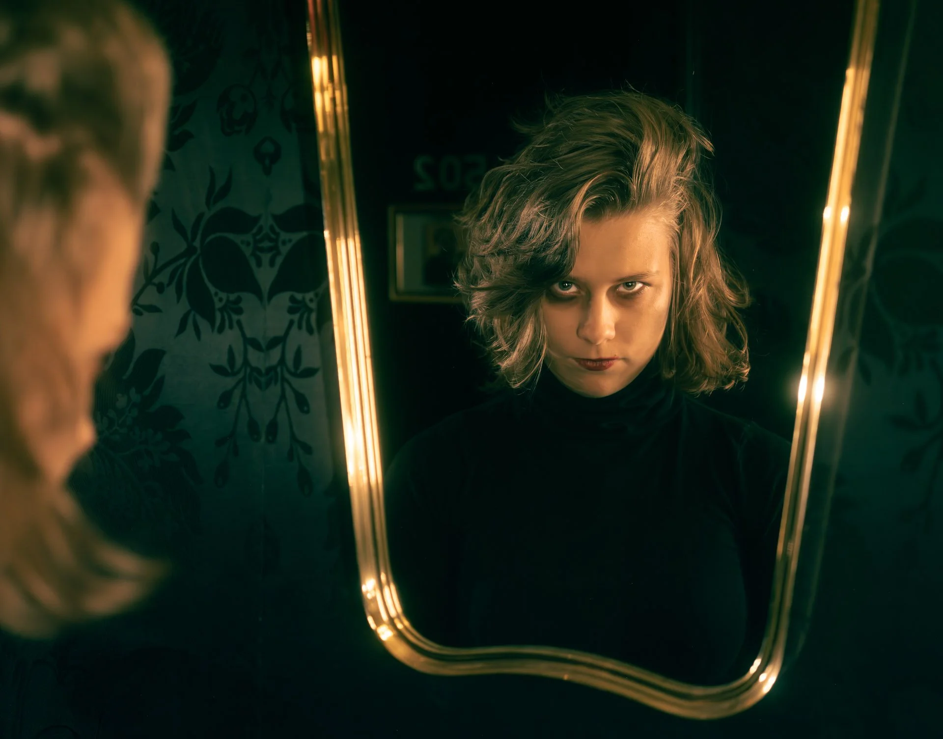 A woman with curly blonde hair looking into a mirror in a dimly lit room with patterned wallpaper. The mirror has a glowing yellow border.