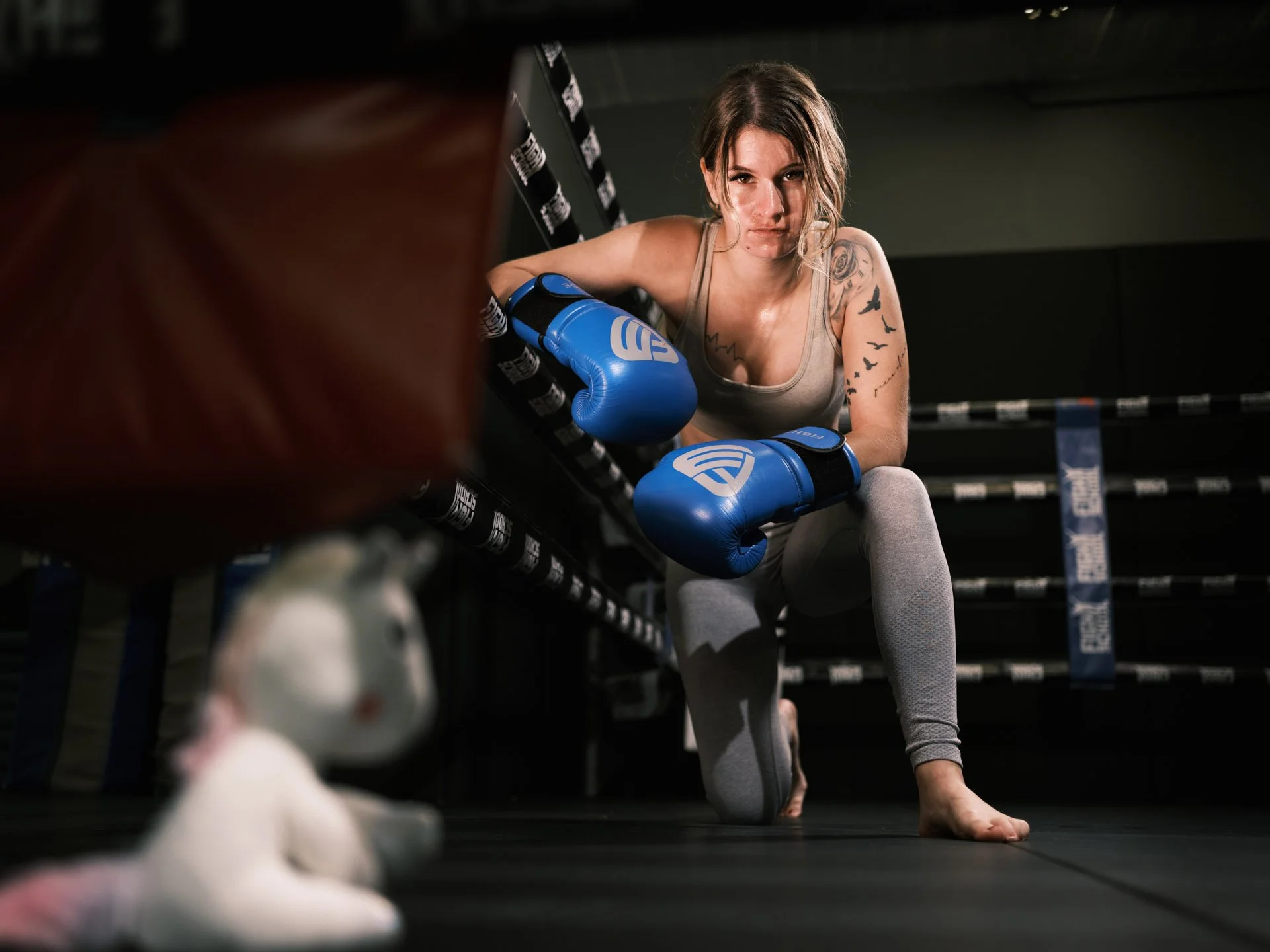 A female boxer wearing grey leggings and a tank top, with boxing gloves, crouching in a boxing ring and looking at the camera.