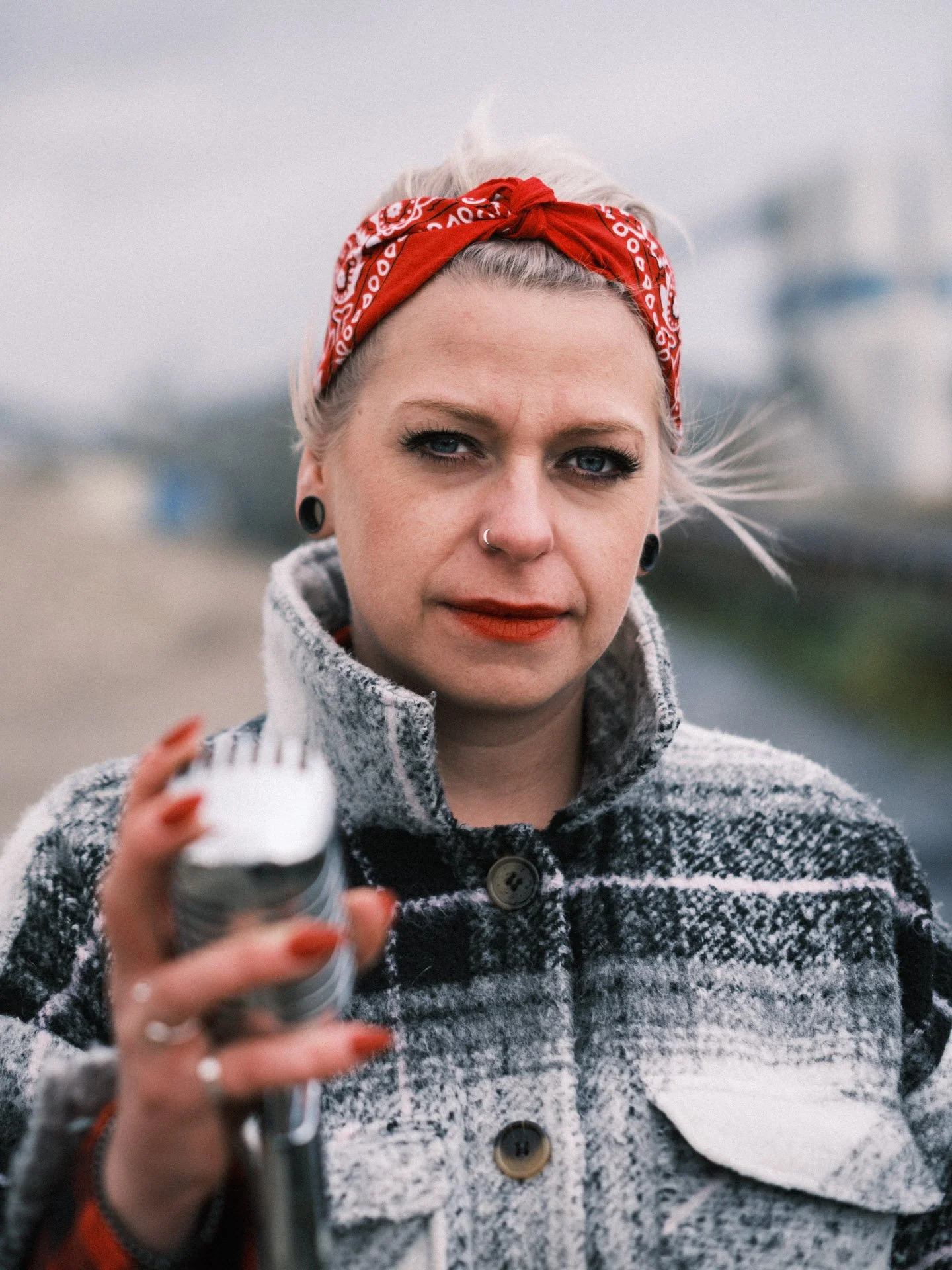 Portrait of a woman with platinum blonde hair, wearing a red bandana, gray and black checkered coat, holding a metal spray bottle, standing outdoors with a blurred overcast background.