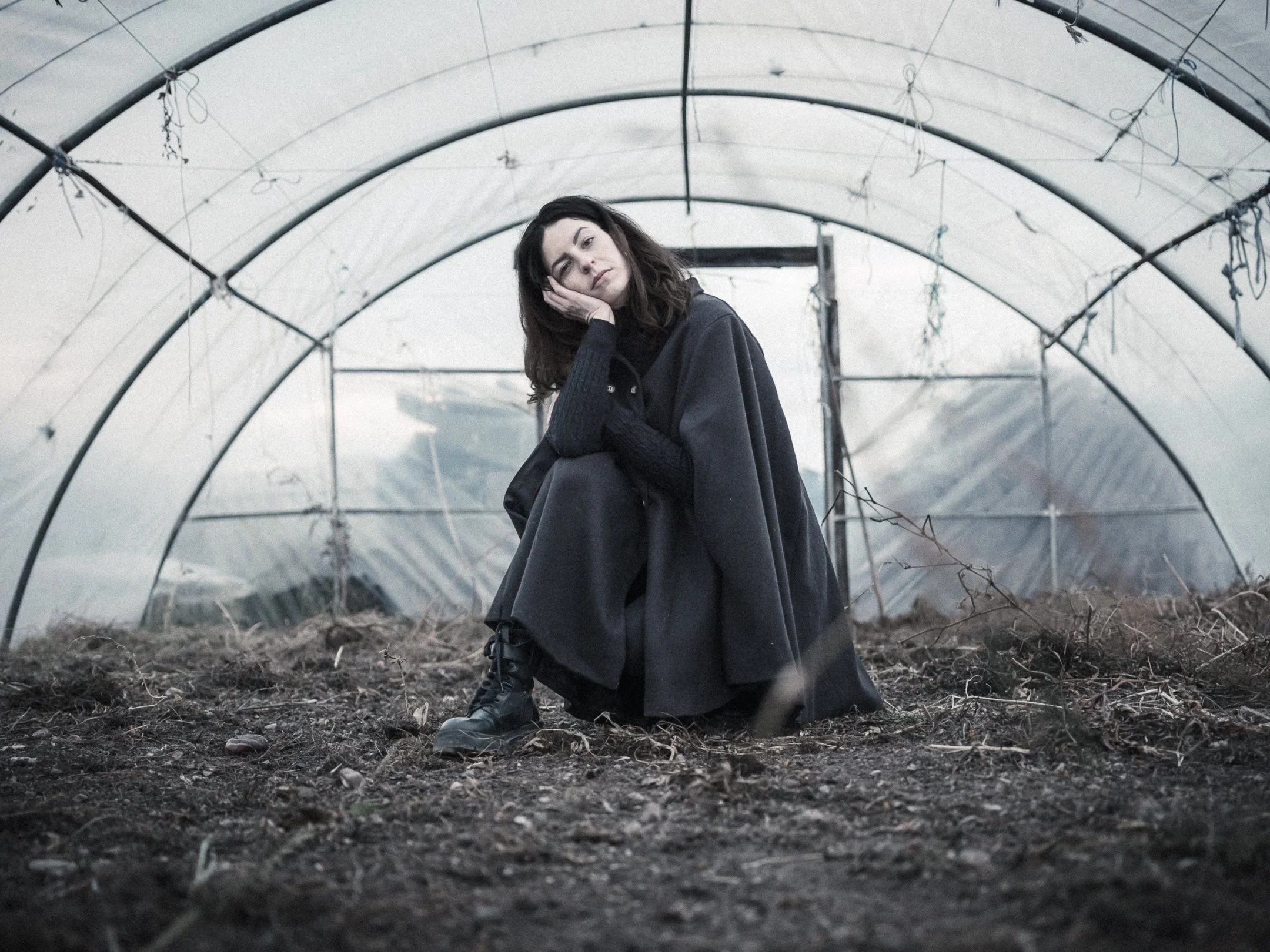 A woman in dark clothing crouches inside a greenhouse with a metal frame, looking at the camera with her head resting on her hand, and the ground is covered in dirt and small rocks.