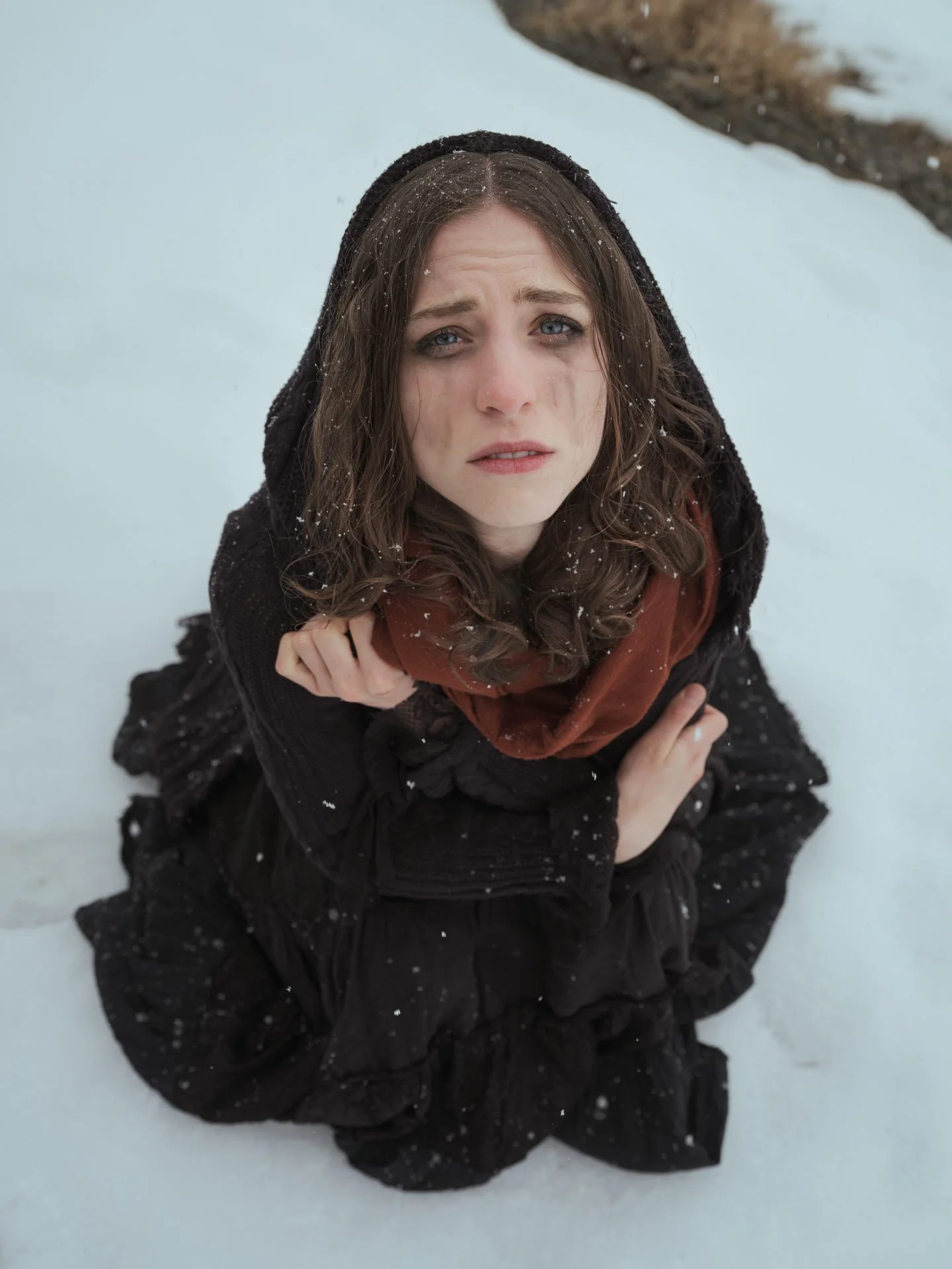 A young woman with blue eyes and brown hair kneeling on snow, looking up at the camera. She is wearing a black hooded coat and a rust-colored scarf. Snowflakes are on her hair and coat.