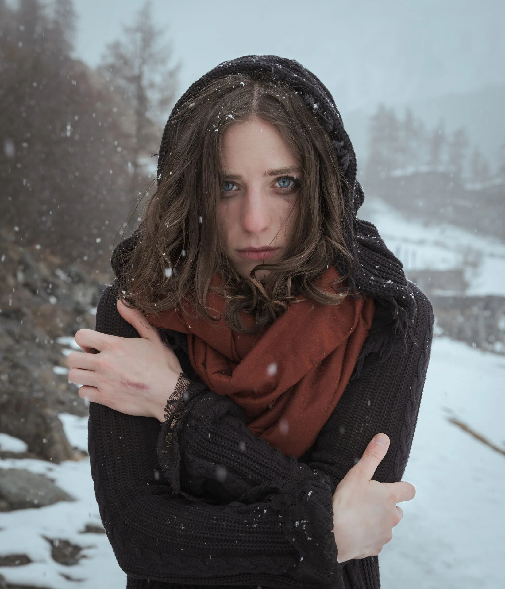 A woman with wet brown hair and blue eyes stands outdoors in a snowy landscape, dressed in a black knit sweater, a rust-colored scarf, and a black beanie, with snowflakes on her hair and clothing.