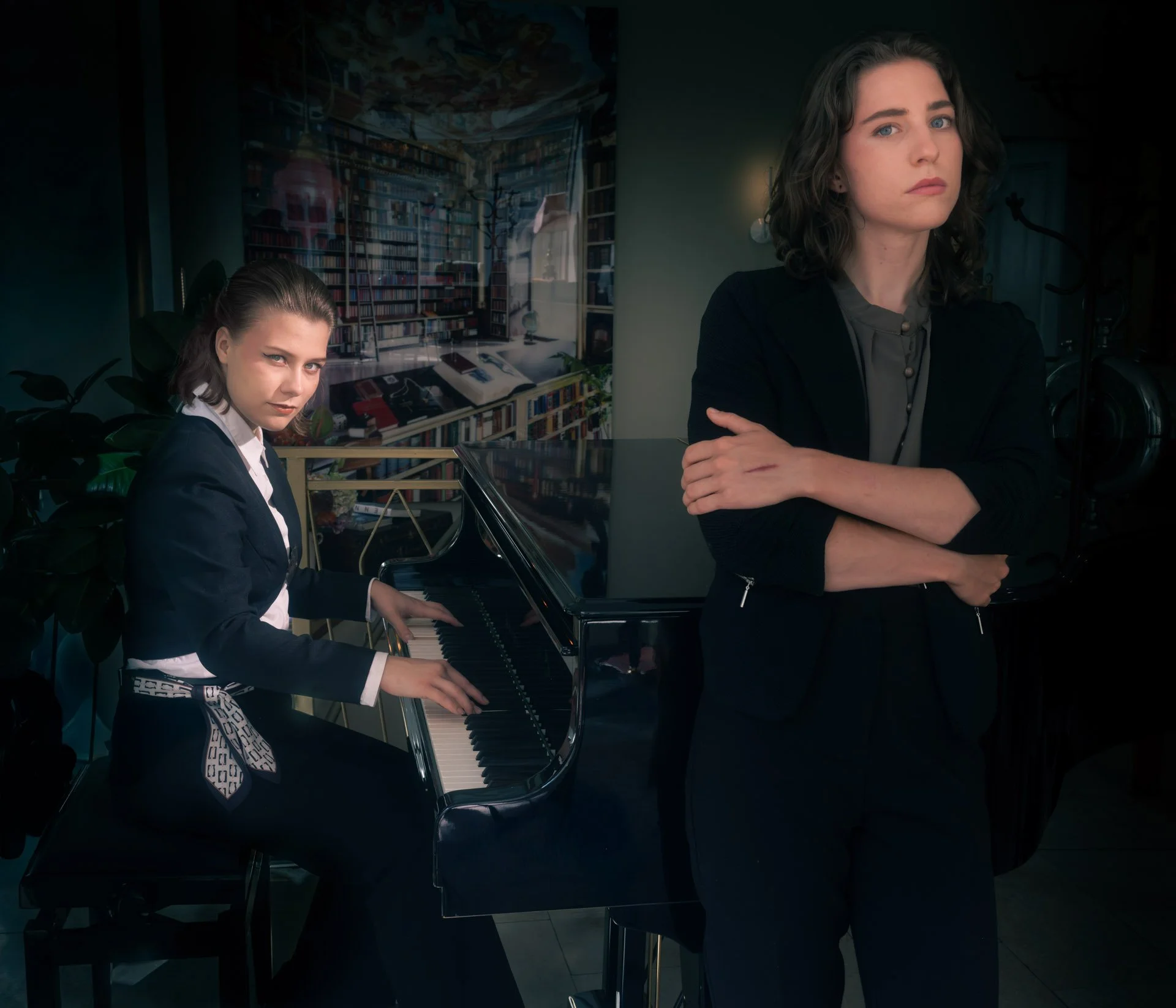 Two women in a room, one playing a black grand piano and the other standing beside it with arms crossed, with a large bookshelf mural on the wall behind them.