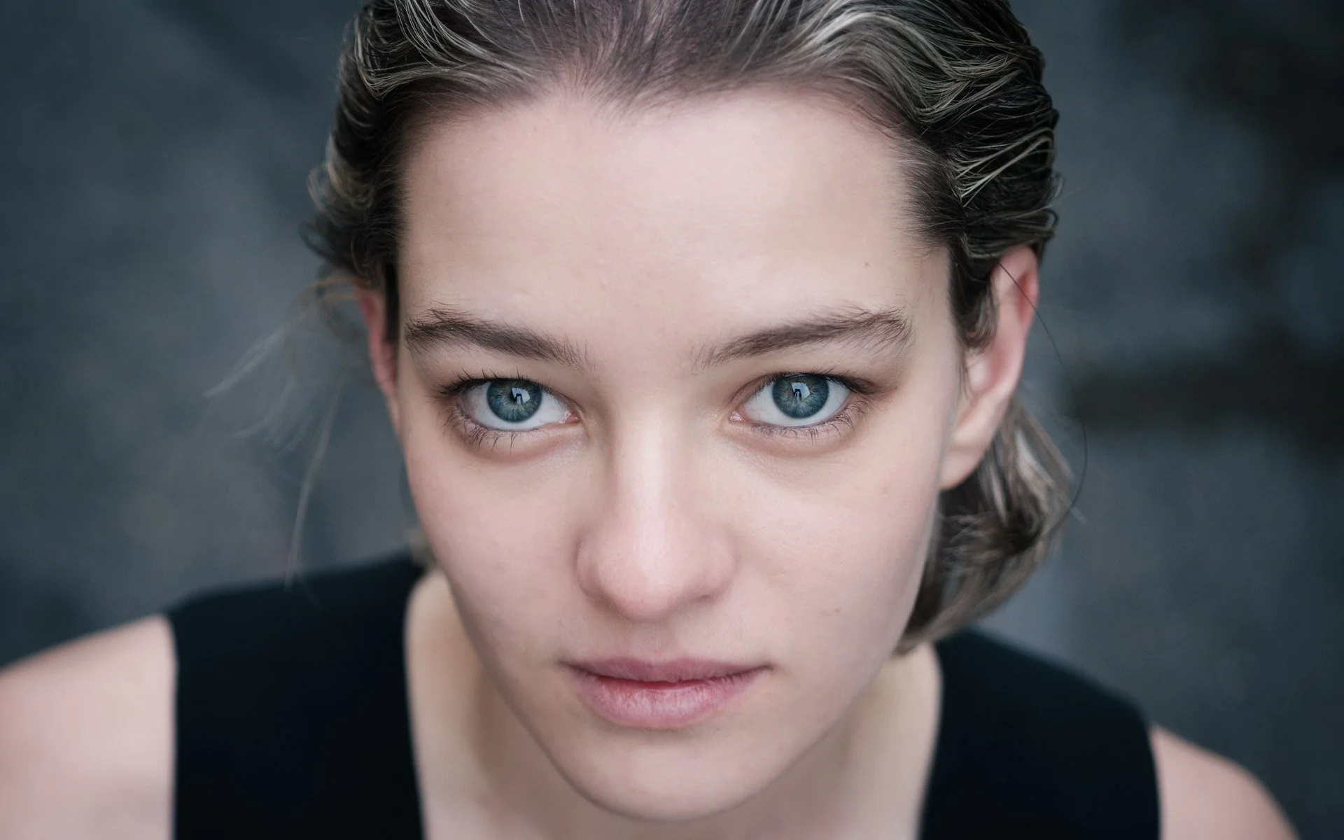 Close-up of a woman with blue eyes and short brown hair, looking directly at the camera against a blurred dark background.