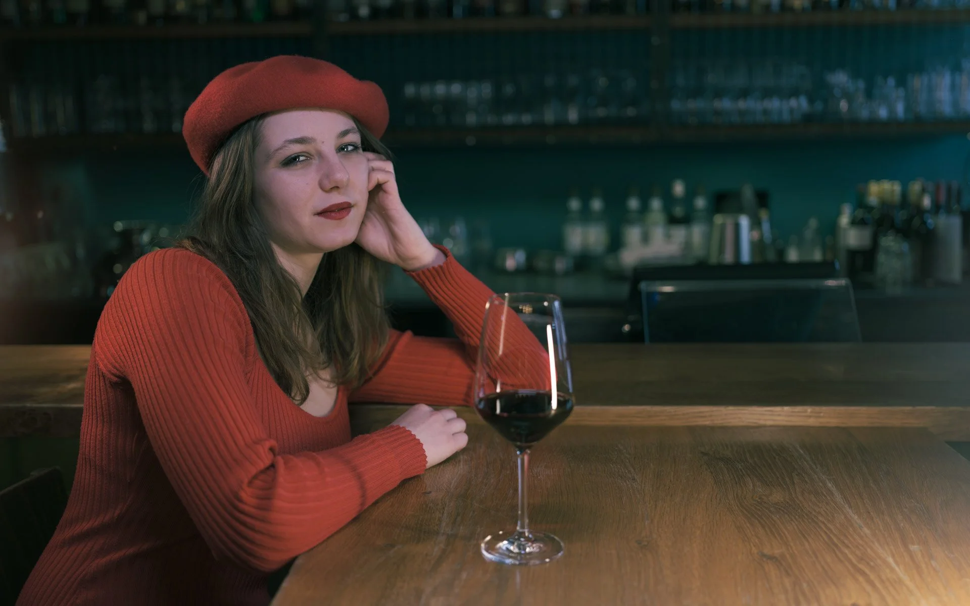 A woman with long brown hair wearing a red beret and red sweater sitting at a bar with a glass of red wine in front of her, in a dimly lit bar or restaurant.