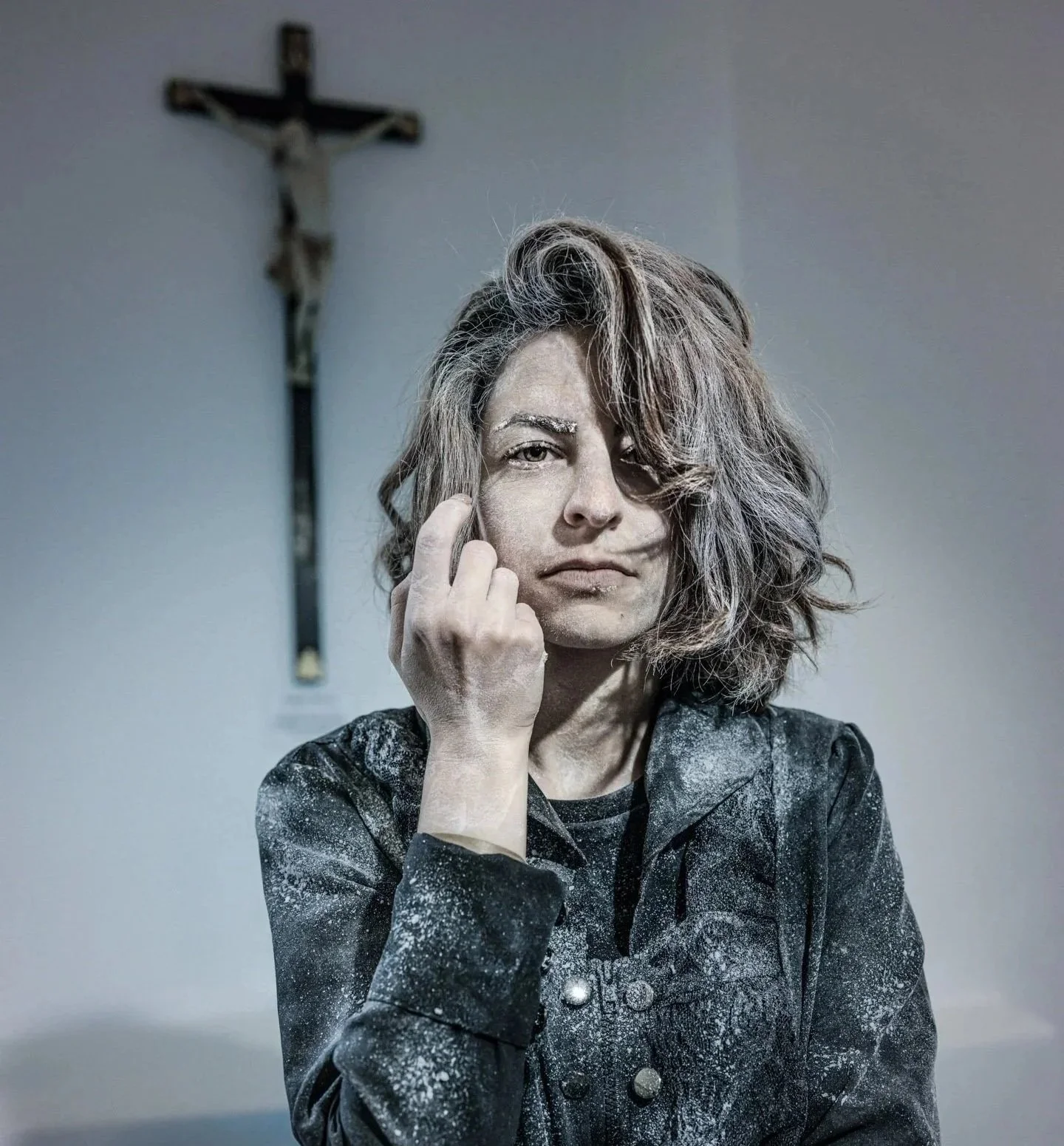 A woman with wavy hair and a serious expression, with her middle finger raised, standing in front of a crucifix on a plain wall.