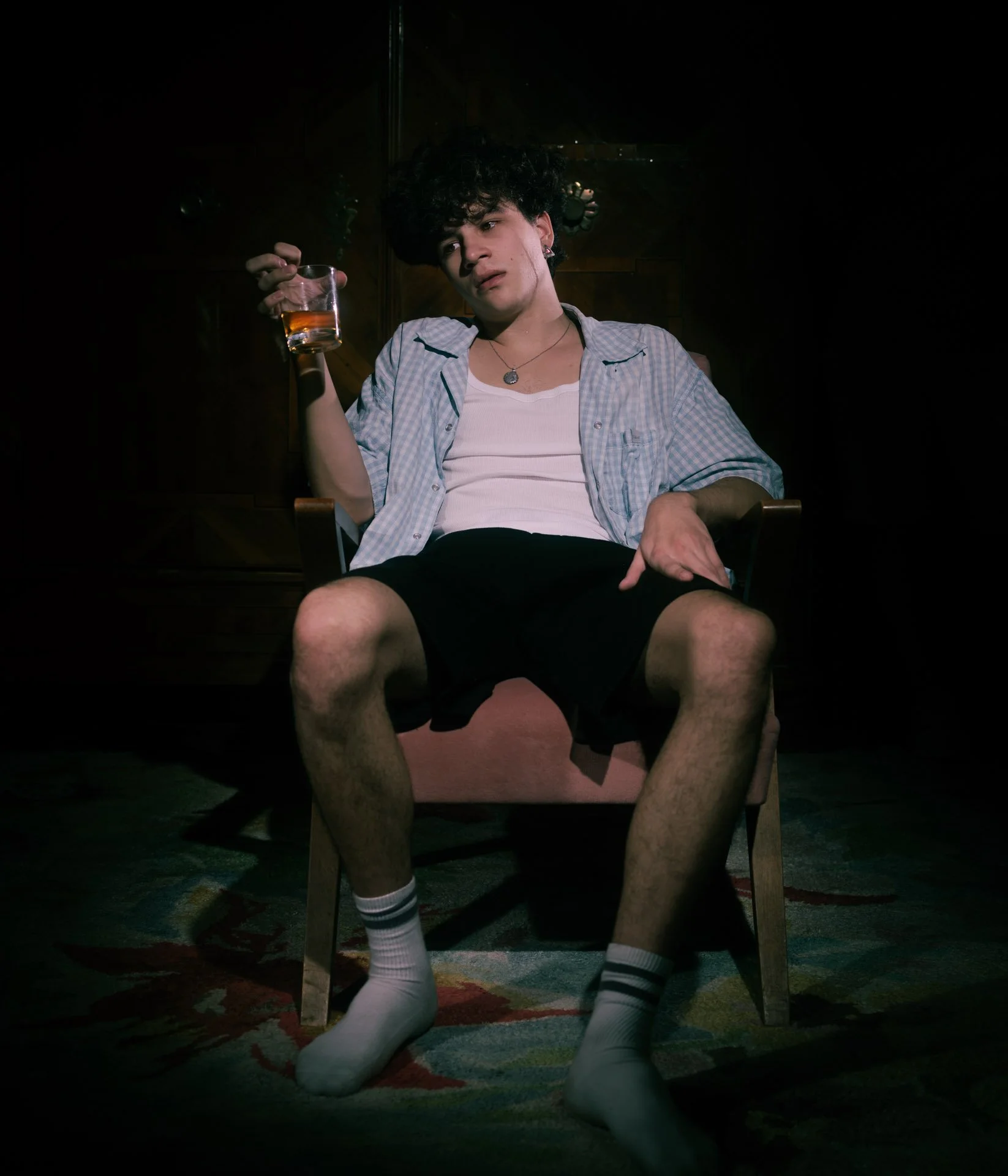 Young man with curly hair sitting in a wooden chair in a dark room, holding a glass with amber-colored liquid in his right hand, wearing a checked shirt over a white tank top, black shorts, and white socks with black stripes.