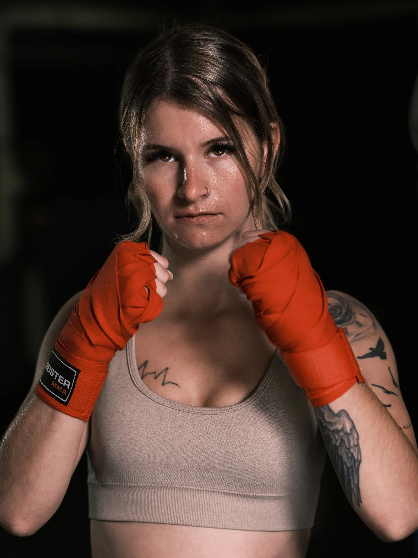 A female boxer in a fighting stance, wearing a beige sports bra and red boxing gloves, with tattoos on her arms and a serious expression.