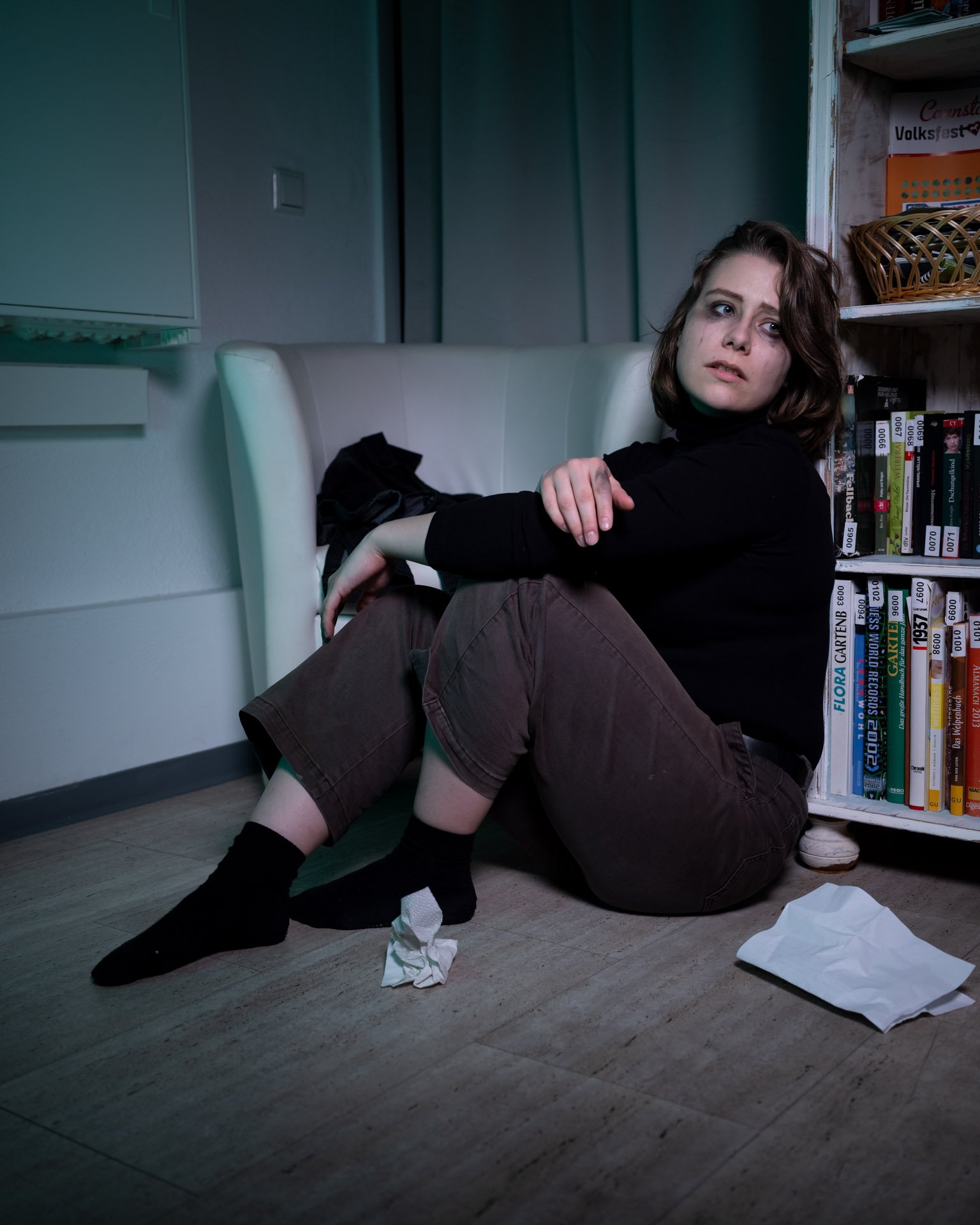 A young woman sitting on the floor next to a bookshelf and a white armchair, with tissues on the floor, looking distressed or upset.
