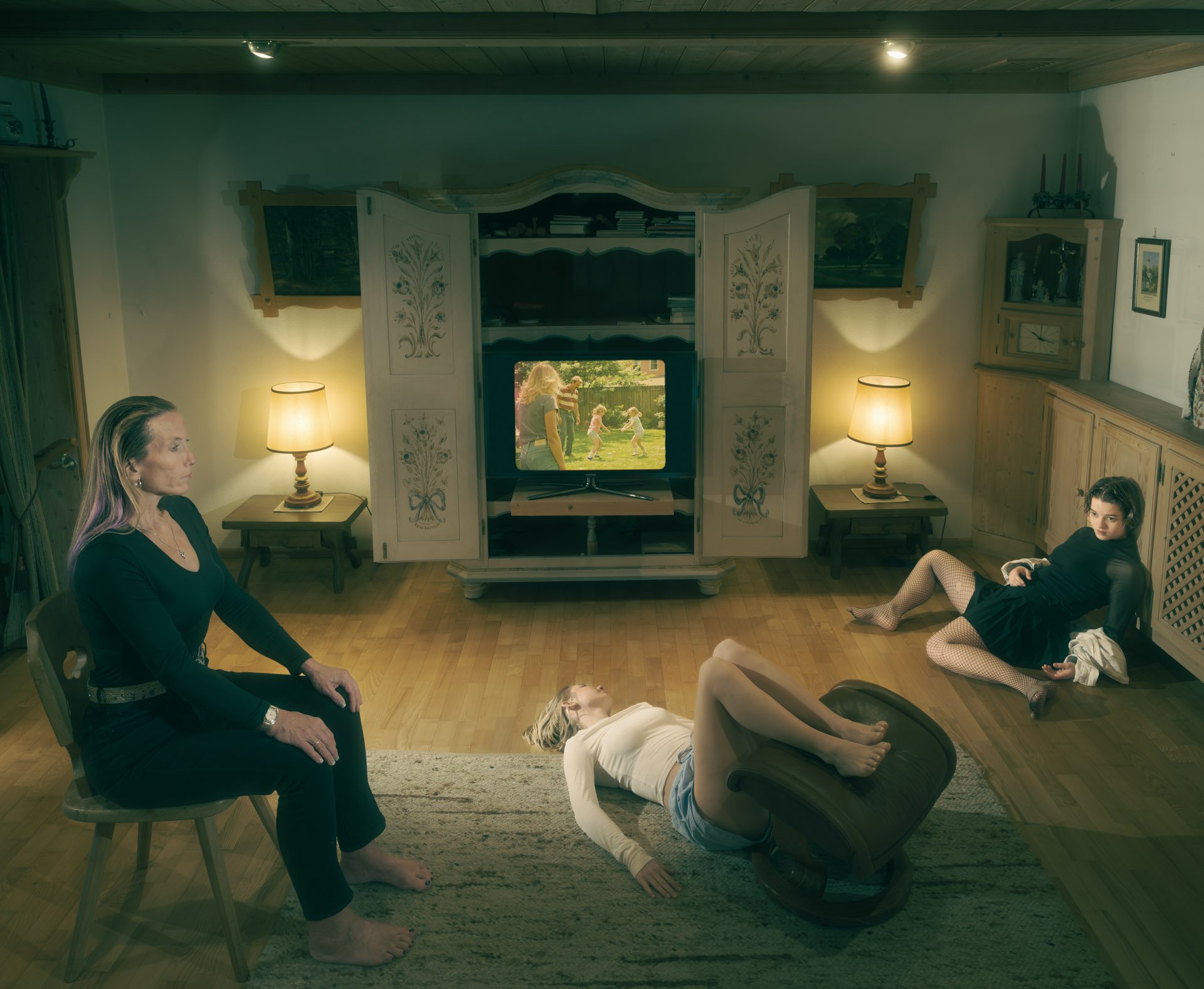 A living room with five people in various relaxed poses. Two women are seated, one with purple hair, both dressed in black. Two women are lying on the floor, one on a patterned rug and one on a chair, with the woman on the floor looking up and the woman in the chair wearing fishnet stockings. The fifth person is a woman sitting on a chair. There is a white bookshelf with hand-painted floral designs, a TV displaying children playing outside, and warm yellow lamps illuminating the room.