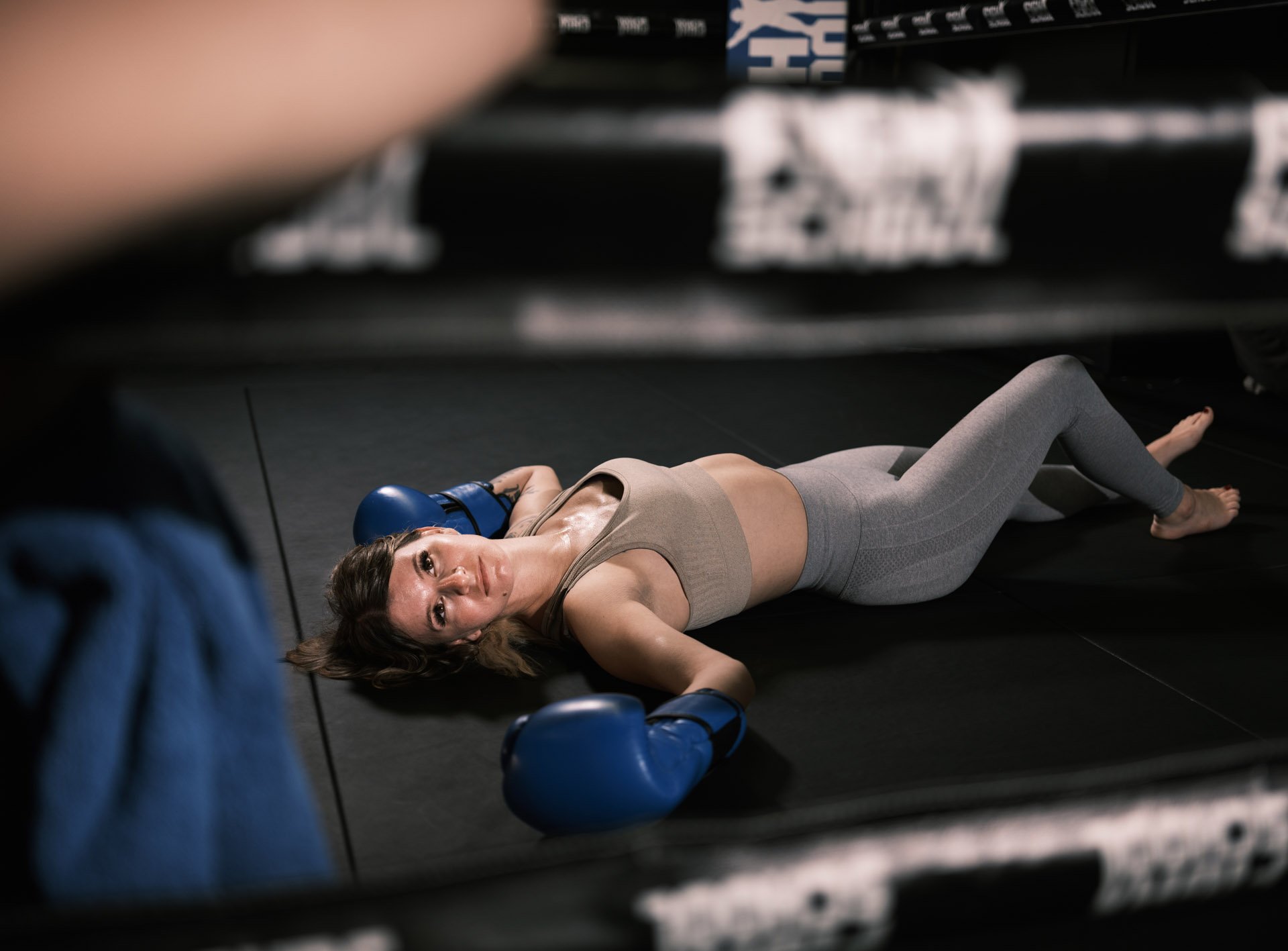 A fit woman lying on the floor of a gym, wearing gray workout leggings, a beige sports bra, and boxing gloves, with a blue boxing helmet, looking relaxed and gazing at the camera.