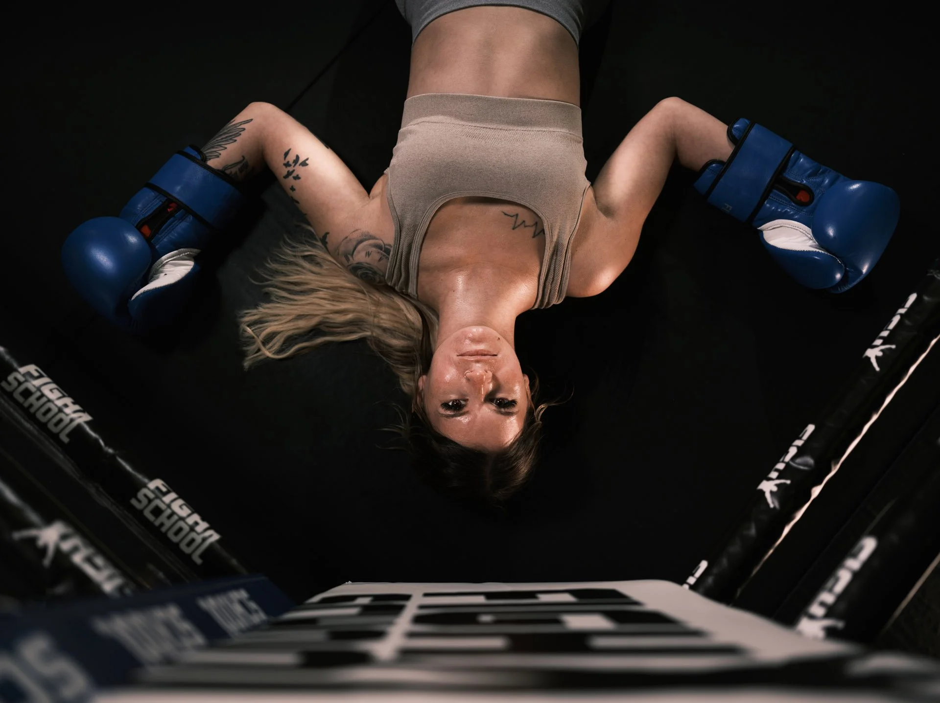 Woman lying on her back inside a boxing ring wearing blue boxing gloves and athletic clothing.