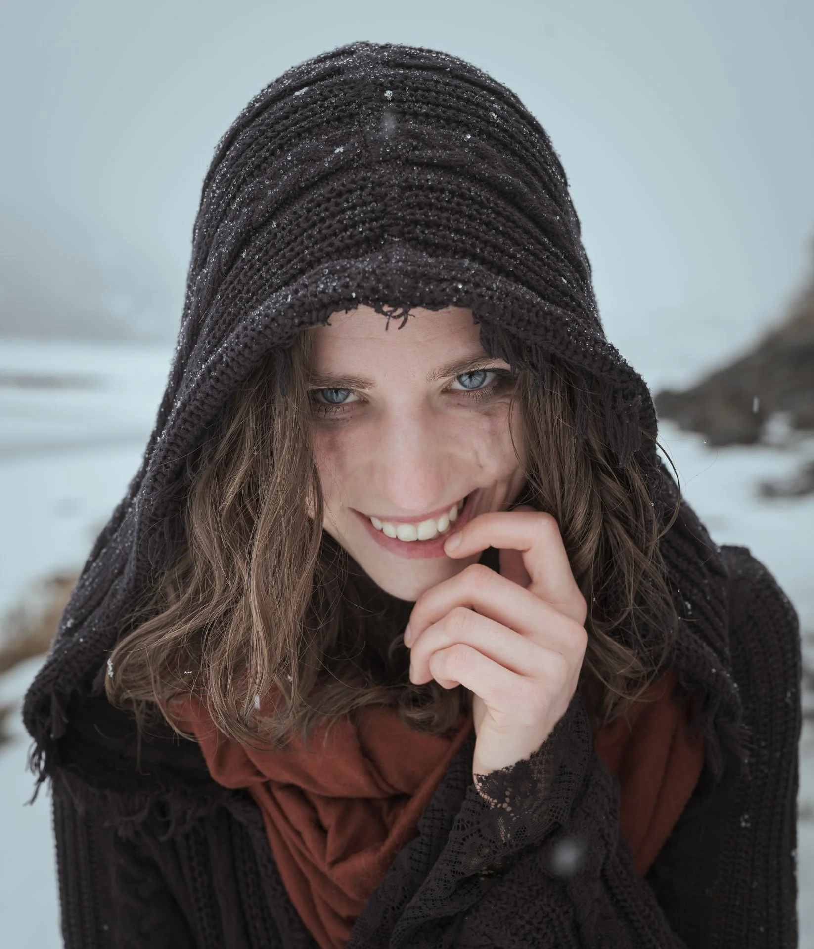 A woman with light brown hair and blue eyes smiling, wearing a dark knitted hooded shawl with snow or ice droplets on it, in a snowy outdoor environment
