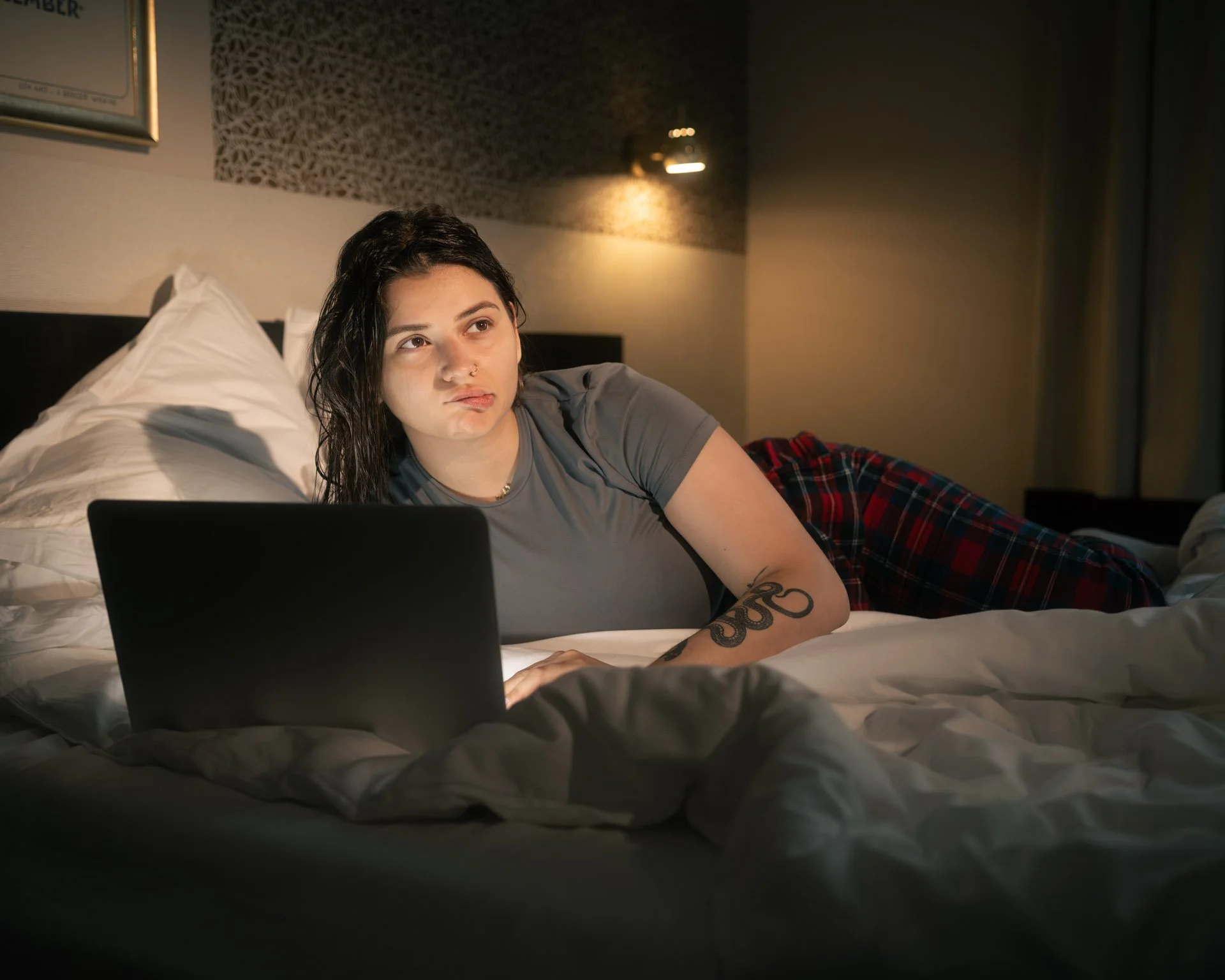 A young woman with dark hair and tattoos lying on her side on a bed, using a laptop. She looks thoughtful or concerned in a hotel room at night.