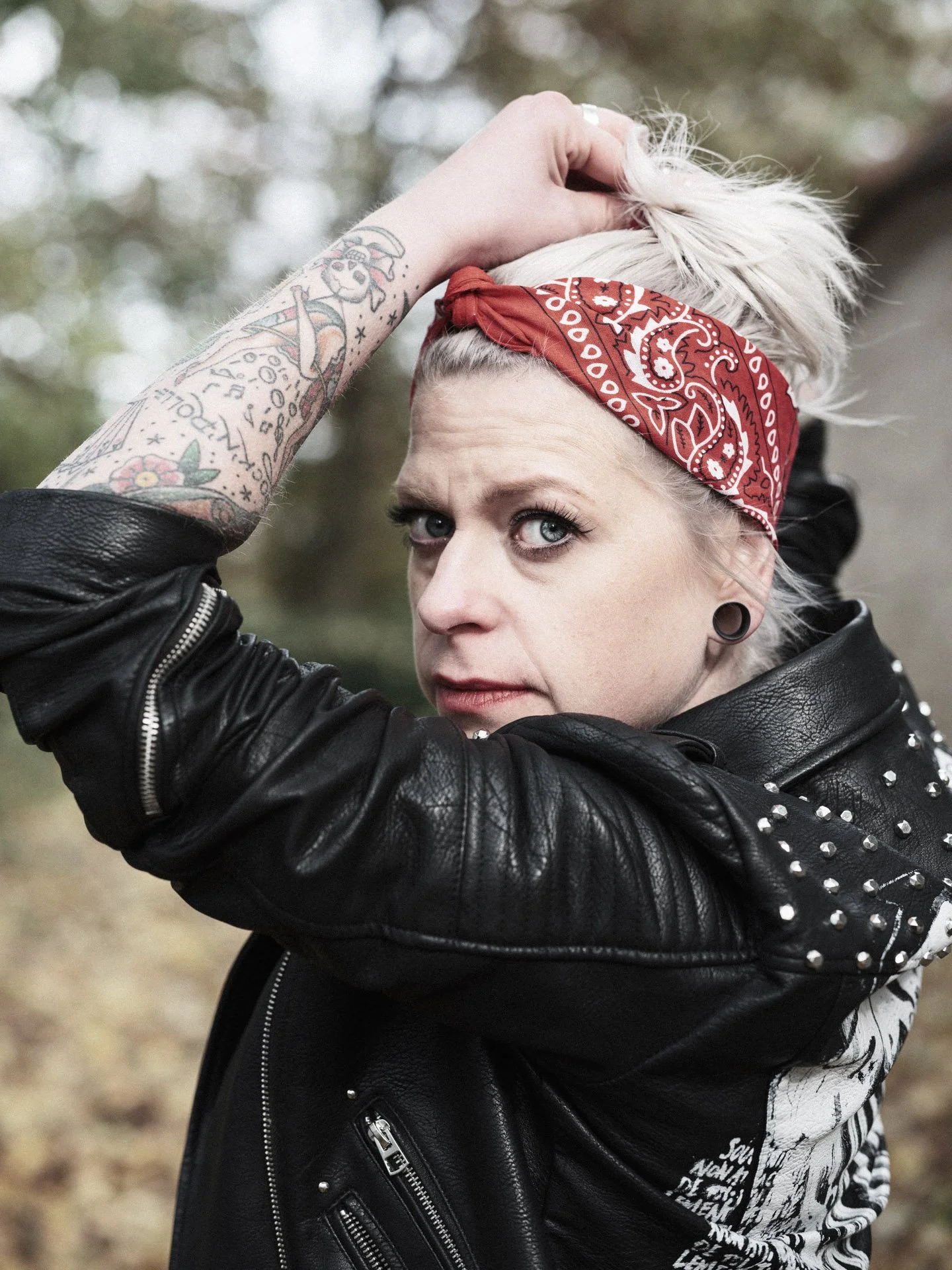 A woman with blonde hair tied back with a red bandana, wearing a black leather jacket with studs, posing outdoors and looking at the camera.
