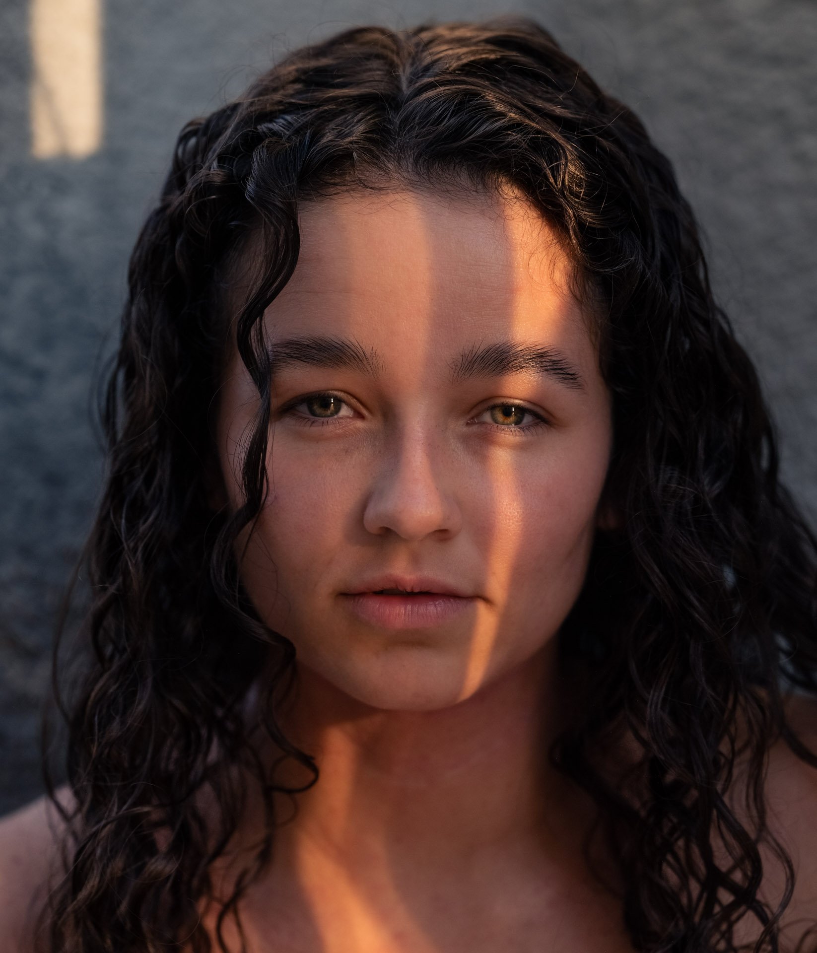 Close-up of a woman with curly dark hair, light complexion, and green eyes, with sunset light casting a shadow on her face.