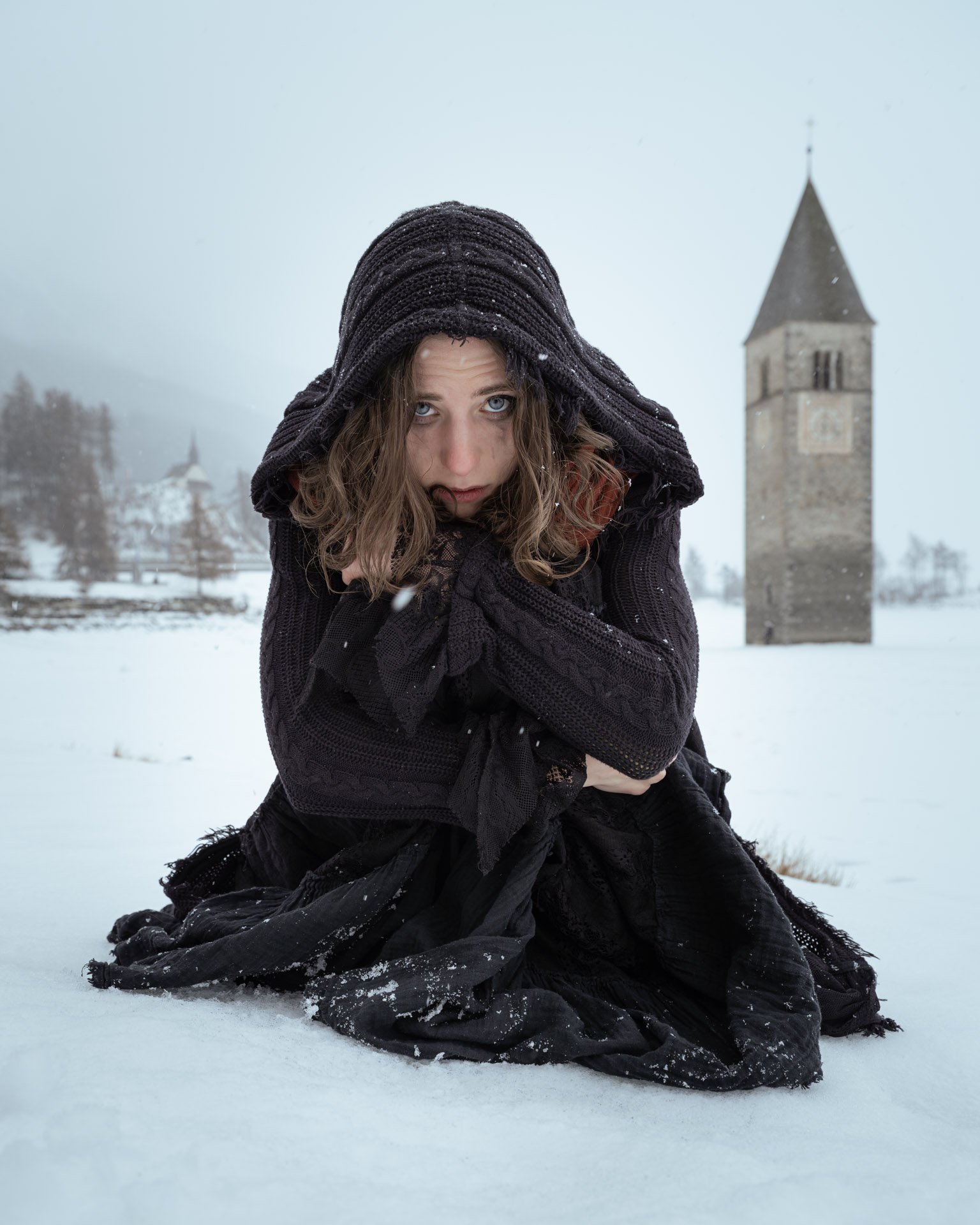 A person in a black hooded cloak kneeling in snow with a church in the background.