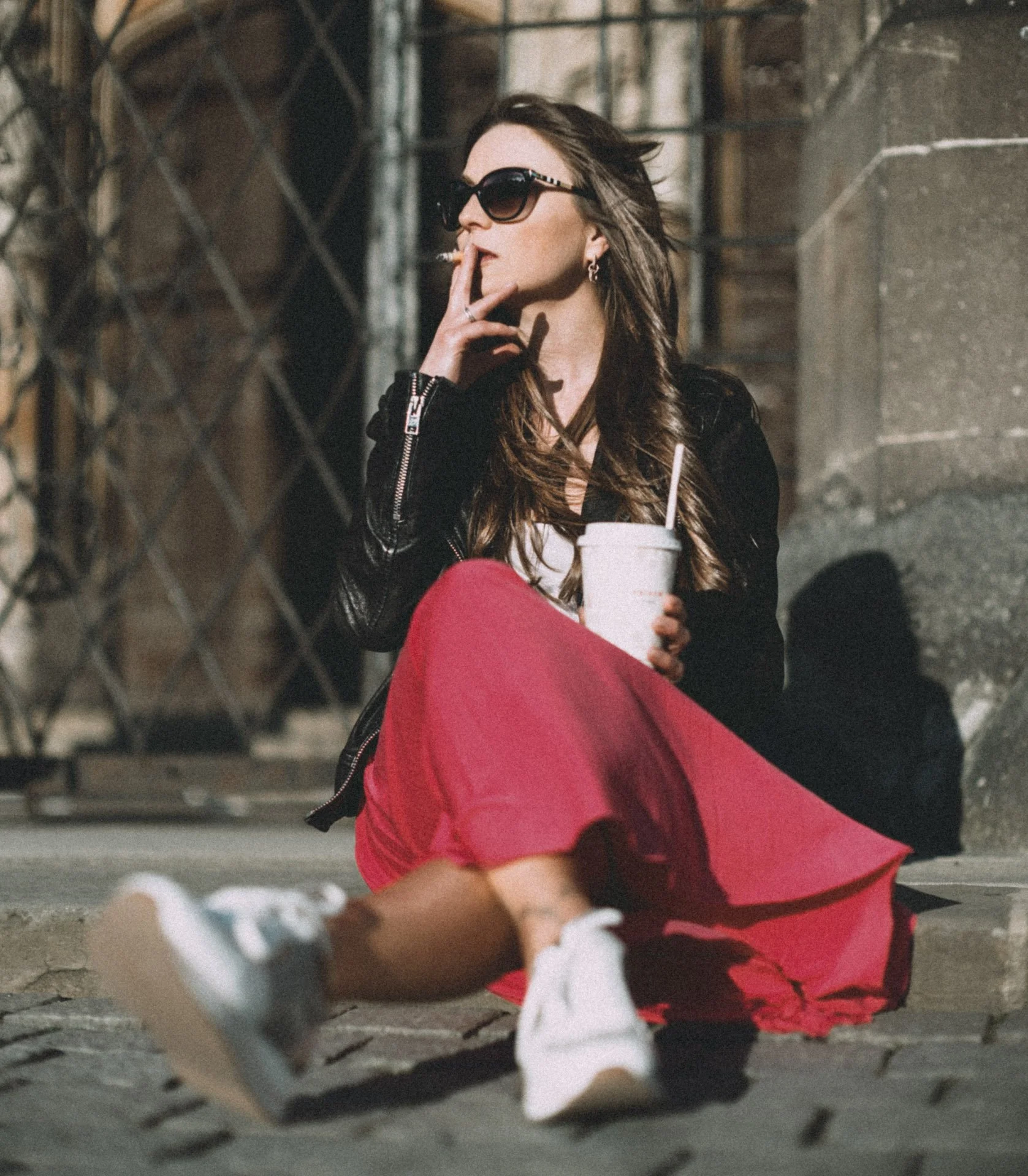Woman with long hair, sunglasses, black leather jacket, white shirt, and red skirt sitting on a city sidewalk against a brick and metal fence, holding a drink and smoking a cigarette.