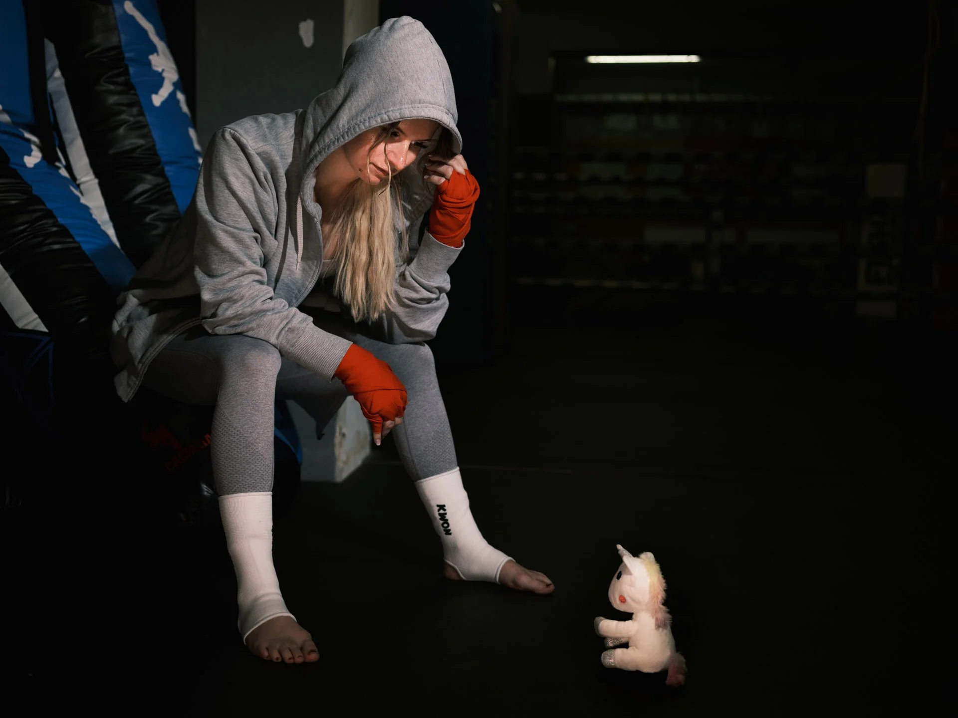 A woman in athletic clothes, with a gray hoodie and leggings, sitting on the floor in a dimly-lit room, looking at a small plush unicorn toy in front of her.
