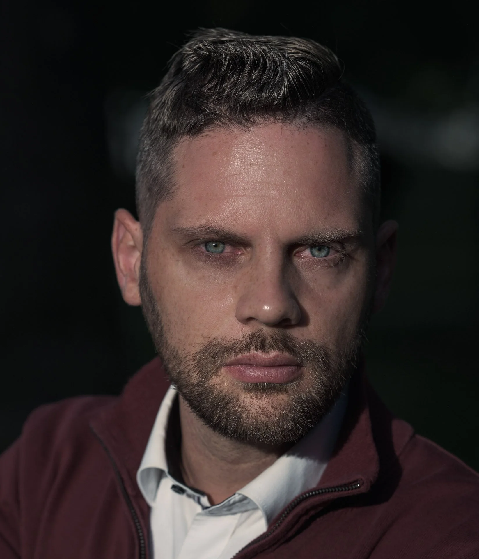 Close-up of a man with short, dark hair, blue eyes, and a beard looking serious, wearing a white shirt and dark red hoodie, outdoors at night.