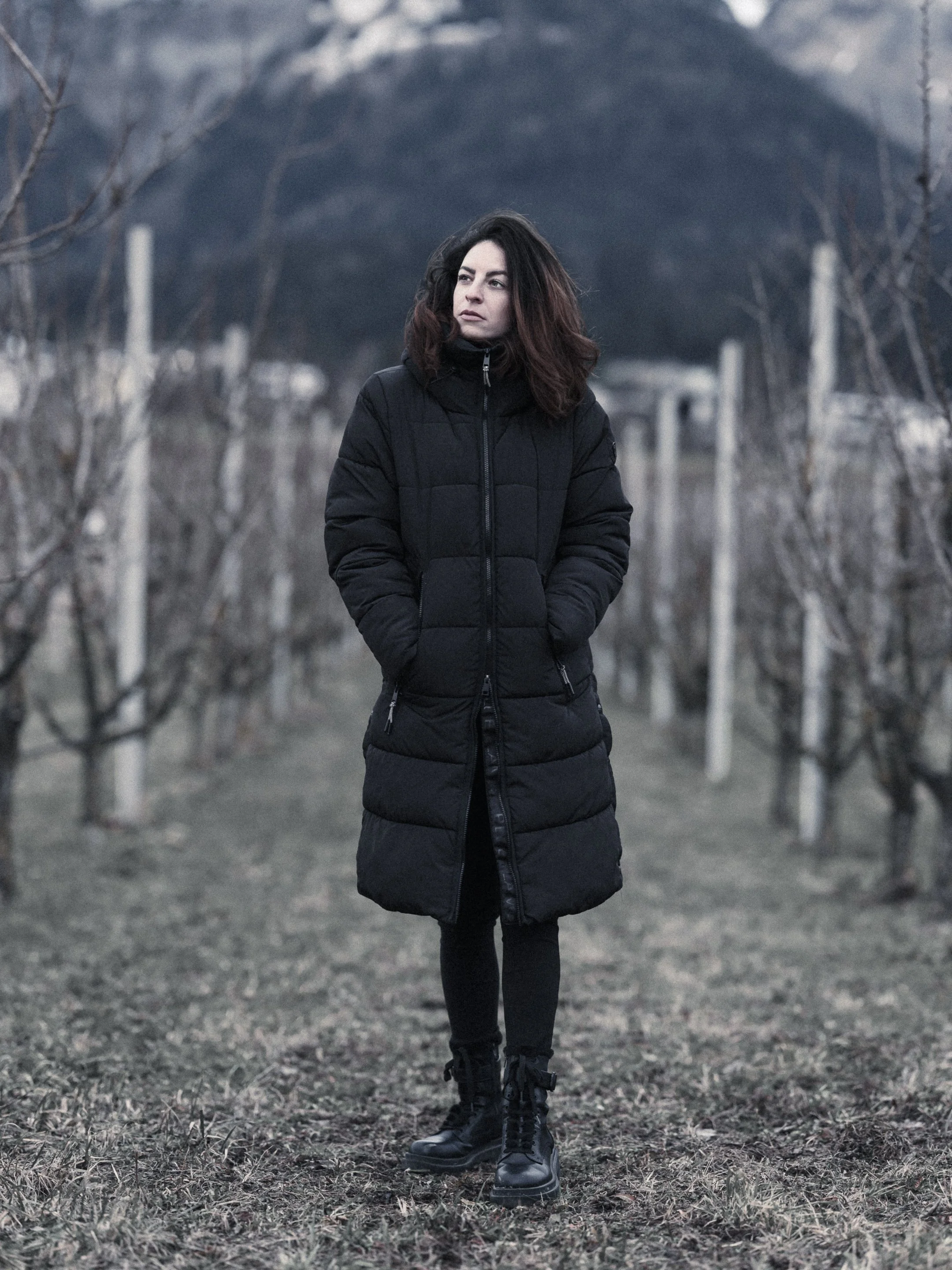 A woman walking outdoors in a winter landscape, wearing a black coat and black boots, with leafless trees and mountains in the background