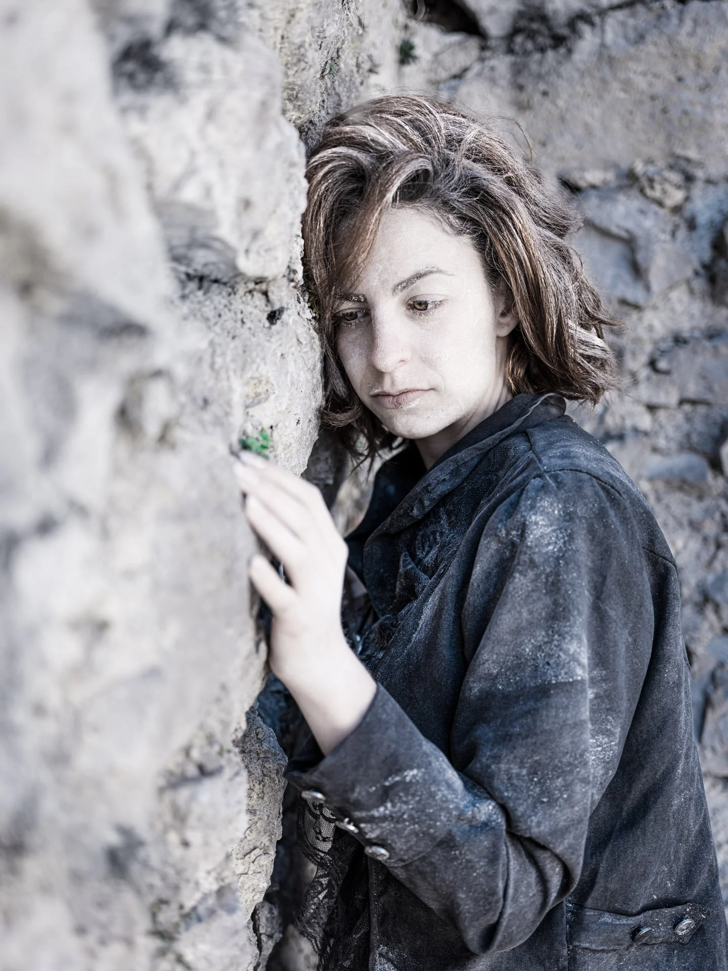 A woman with wavy, shoulder-length brown hair wearing a dark coat, standing close to a rough stone wall, looking pensively to the side.