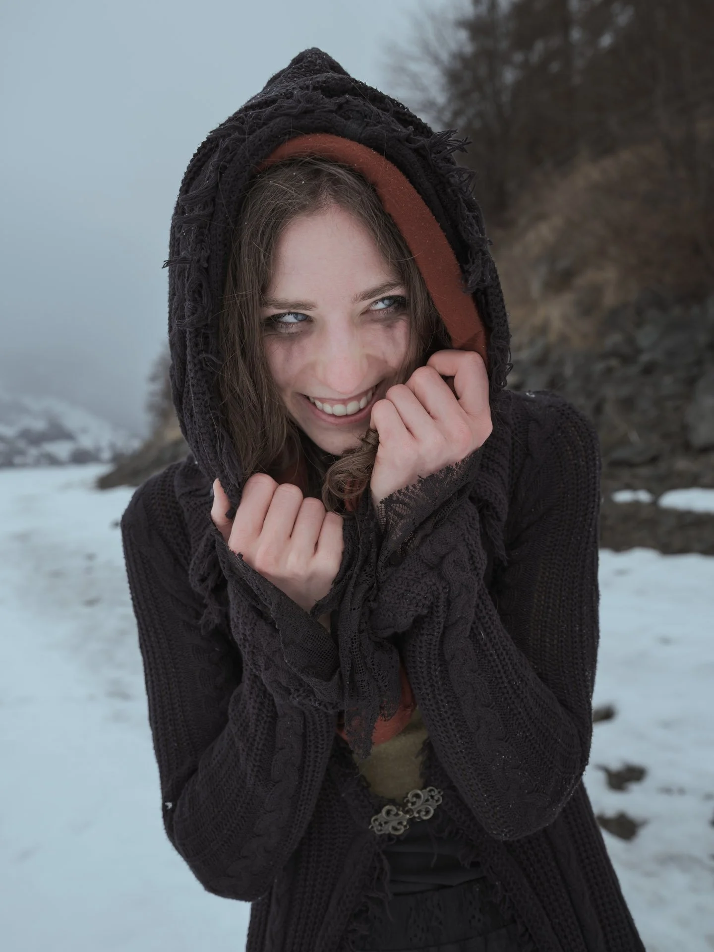 A young woman with brown hair and blue eyes smiling outdoors in a snowy landscape, wearing a black hooded sweater and jewelry.