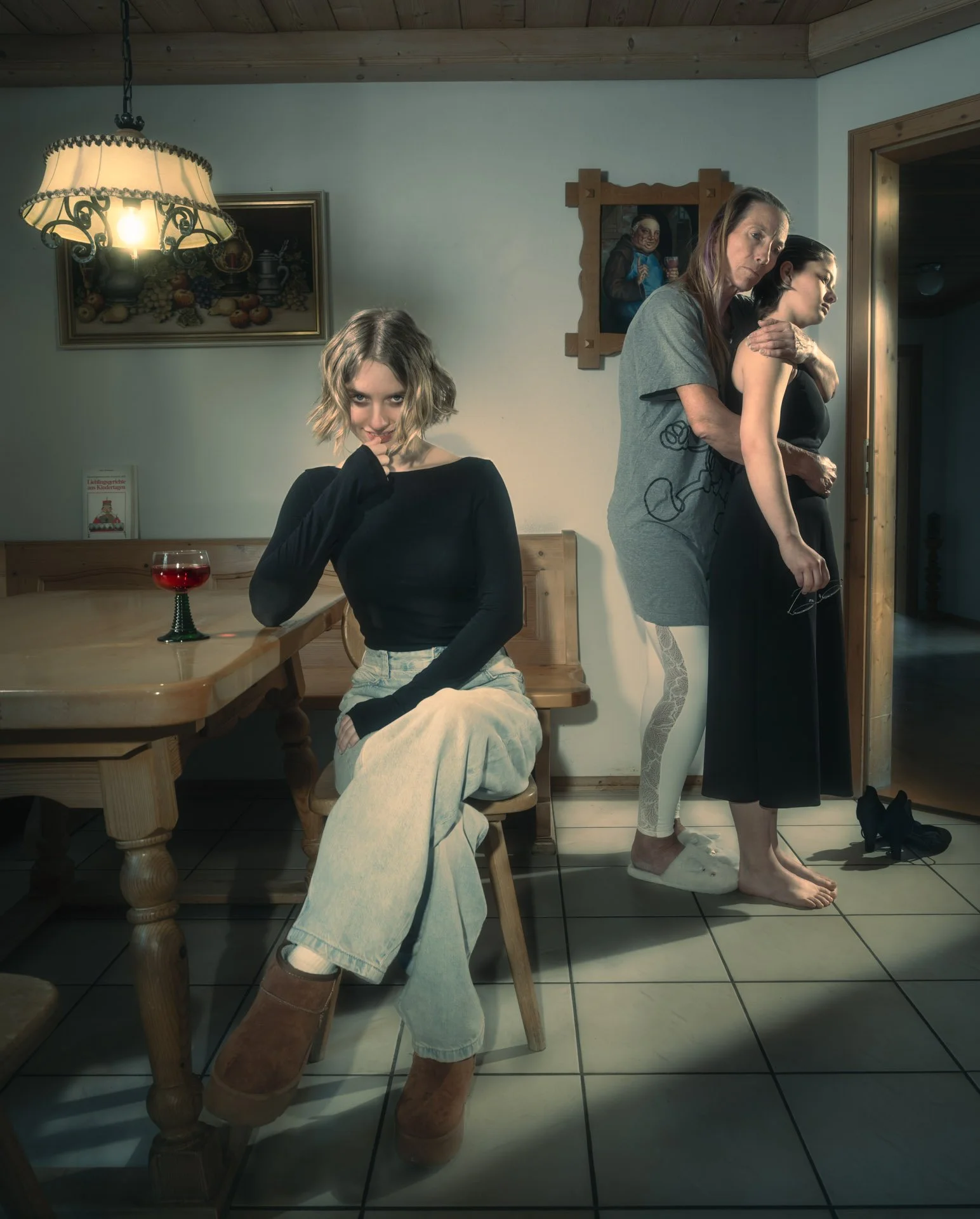 A woman with short blond hair sitting at a wooden table with a glass of red wine, wearing a black long-sleeve shirt and beige pants. Behind her, two women are standing: one with long brown hair, wearing a gray t-shirt and white leggings, seemingly comforting the other woman with black hair in a black dress. The two women standing are barefoot on a tiled floor in a cozy room with wooden accents and paintings on the wall.