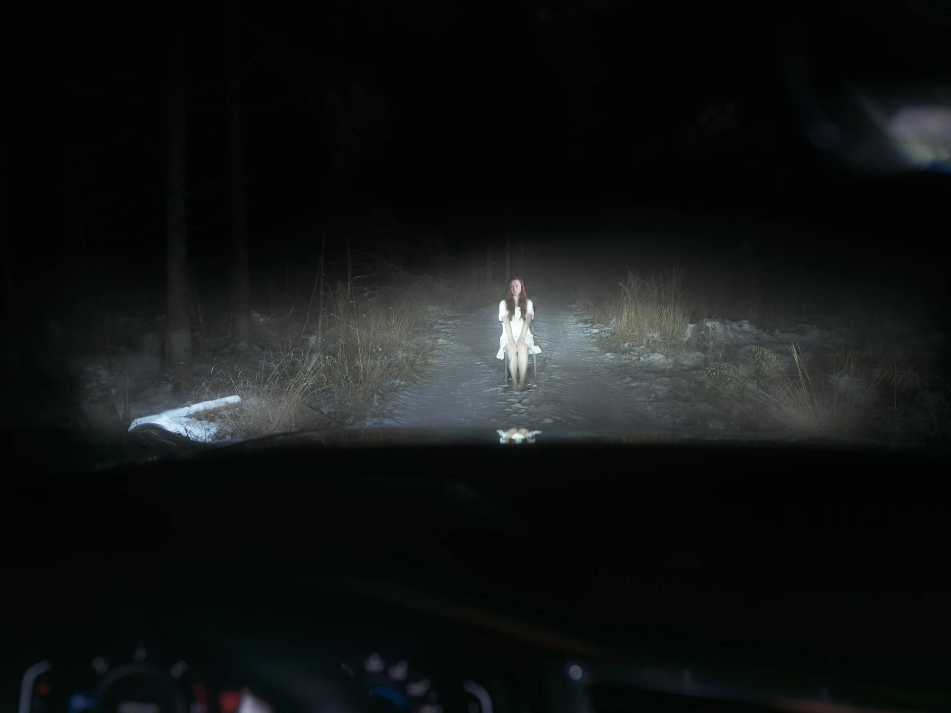 A young woman in a white dress is sitting on a small wooden chair in the middle of a dirt road at night. The surroundings are dark with some trees and grasses visible along the sides of the road.