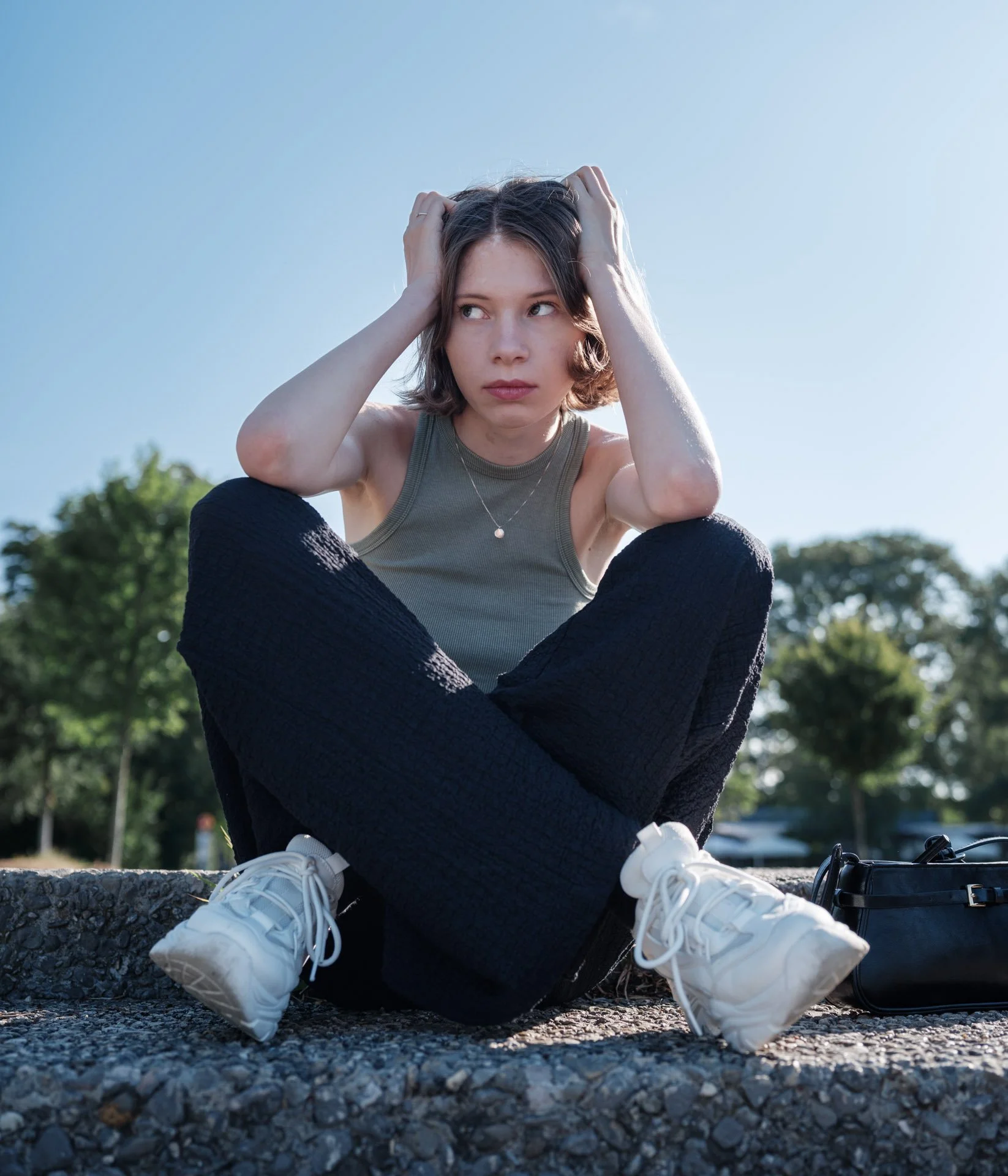 Young woman sitting with her legs crossed outdoors, wearing a beige tank top, black pants, and white sneakers, with a black bag on her side, holding her head with both hands and looking thoughtfully to the side.