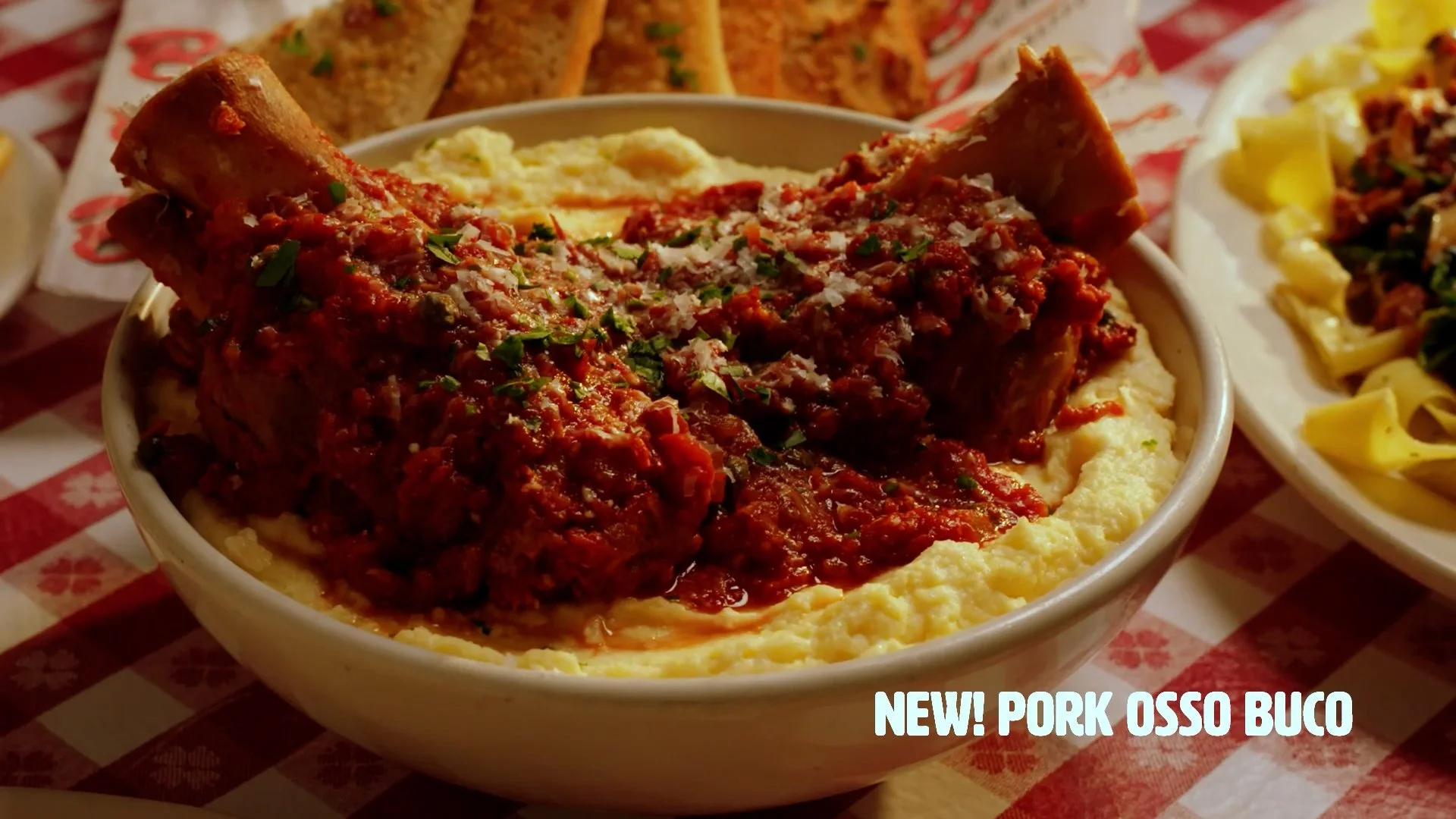 Bowl of mashed potatoes topped with pork ossobuco stew garnished with chopped parsley, surrounded by plates of pasta and bread on a red checkered tablecloth.