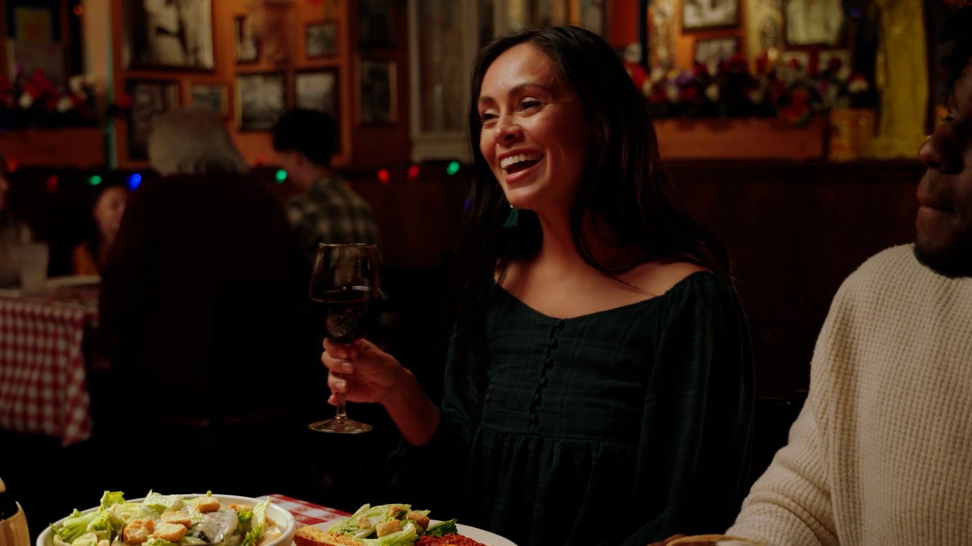 A woman smiling and holding a glass of red wine at a restaurant or party, with a salad and other food on the table in front of her. The background shows a dimly lit, cozy setting with other people dining and festive decorations.