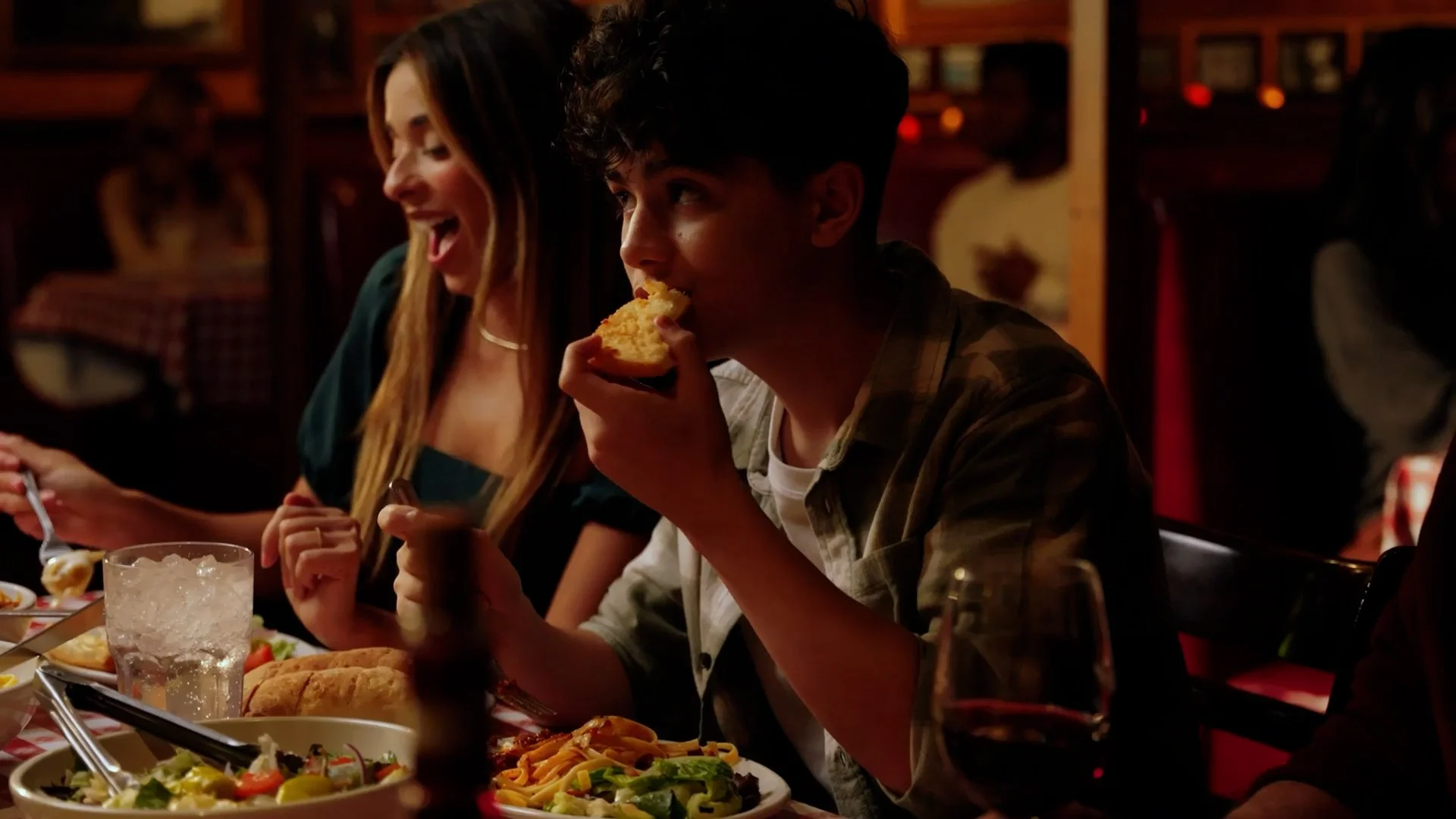 Two young people, a girl with long hair and a boy eating pizza, are sitting at a dinner table in a cozy restaurant with warm lighting. The table has salads, bread, and a glass of water.
