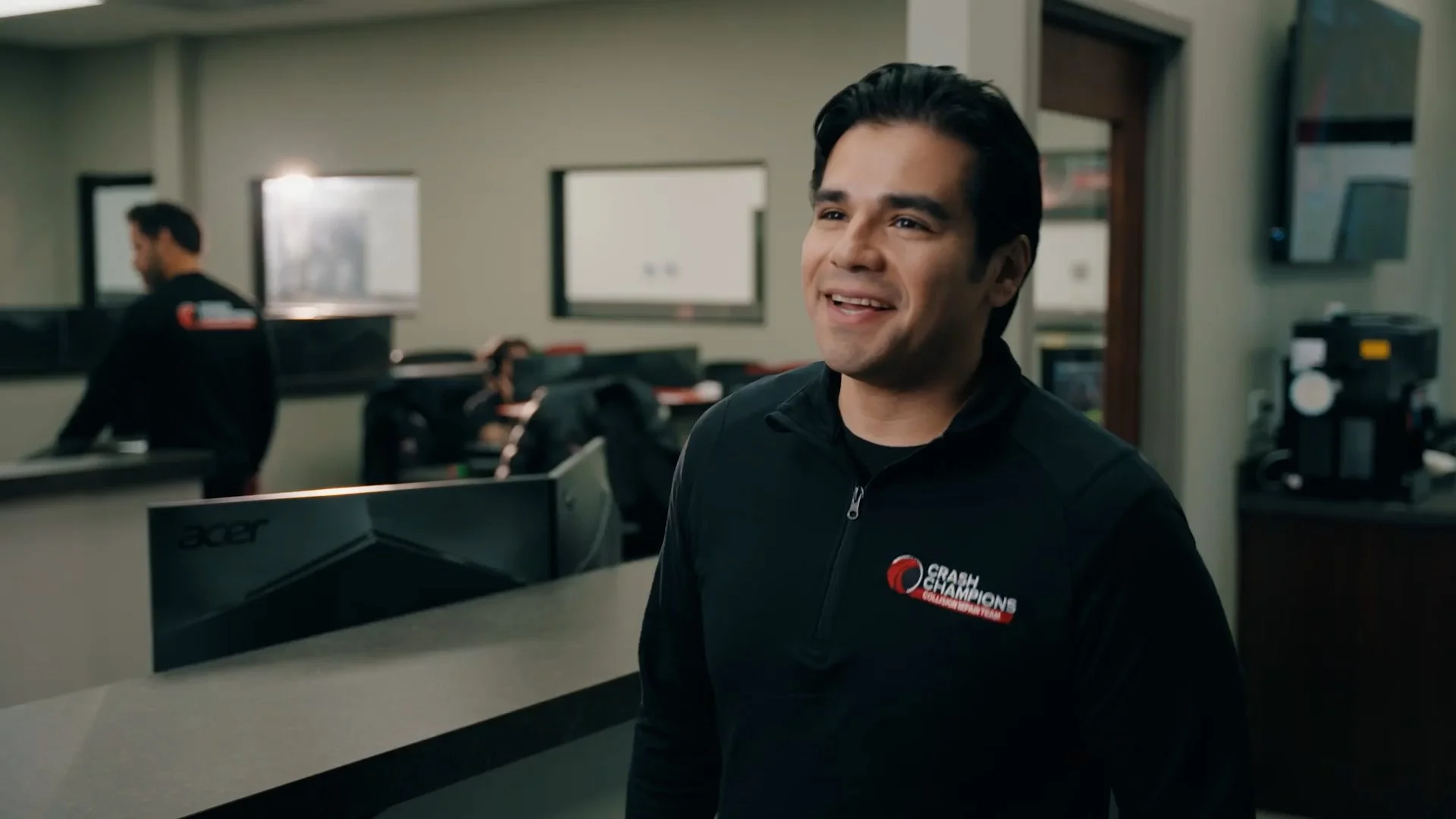 A man smiling in a professional sports team office with a logo on his jacket that reads 'Crash Champions'. The background includes a reception desk, a computer monitor, and other team members.