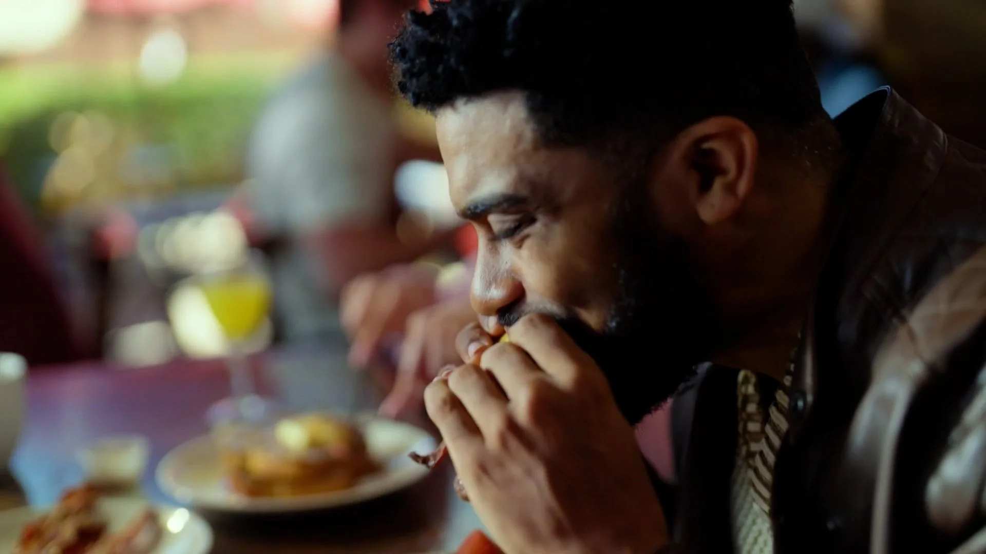 A man with a beard and short curly hair is sitting at a table, holding his hands close to his mouth, appearing to be praying or deeply thinking. There are plates of food and drinks on the table, with other people in the background.